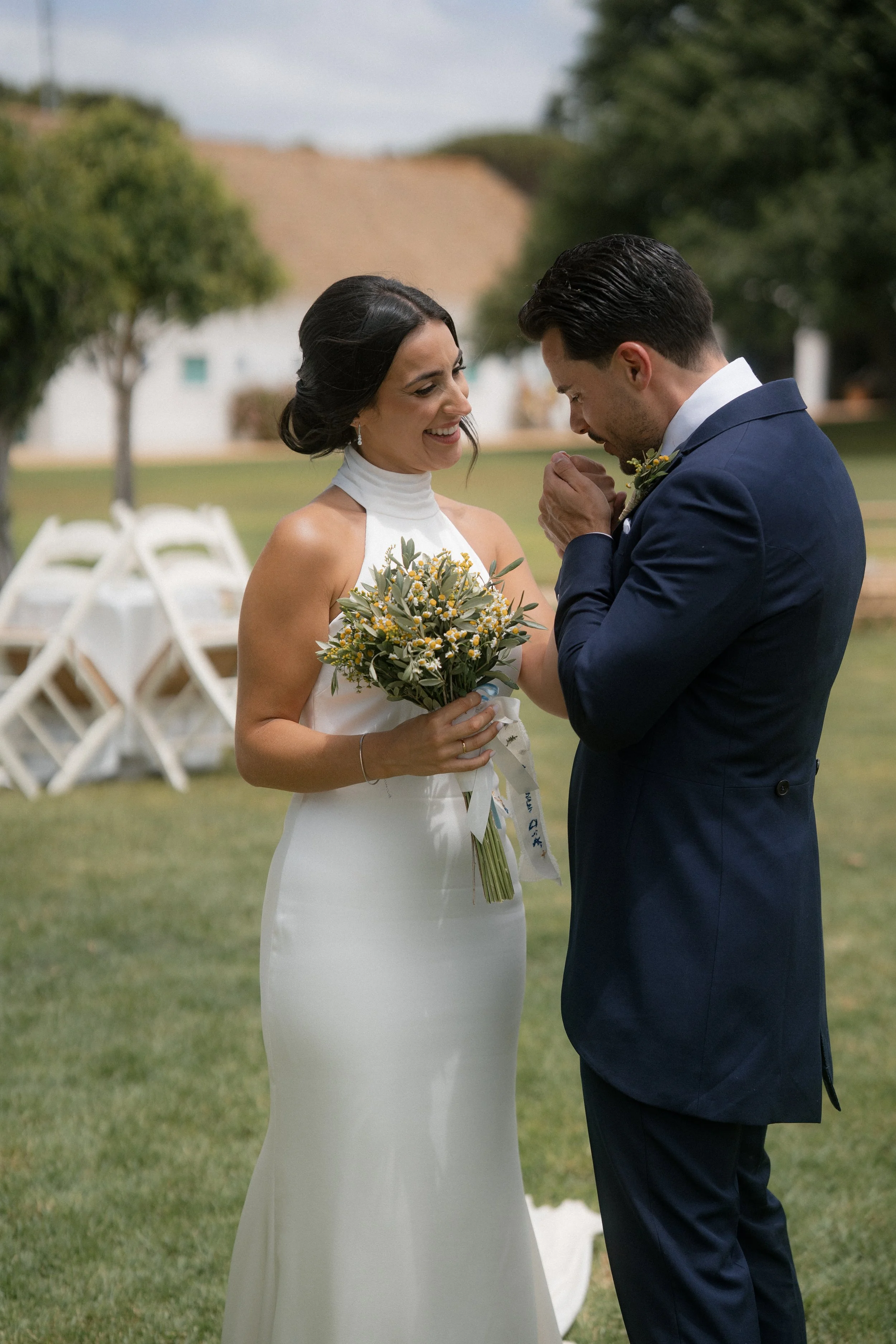 Una pareja de novios en una ceremonia de boda al aire libre, la novia con vestido blanco y el ramo de flores, el novio en traje azul, compartiendo un momento emotivo.