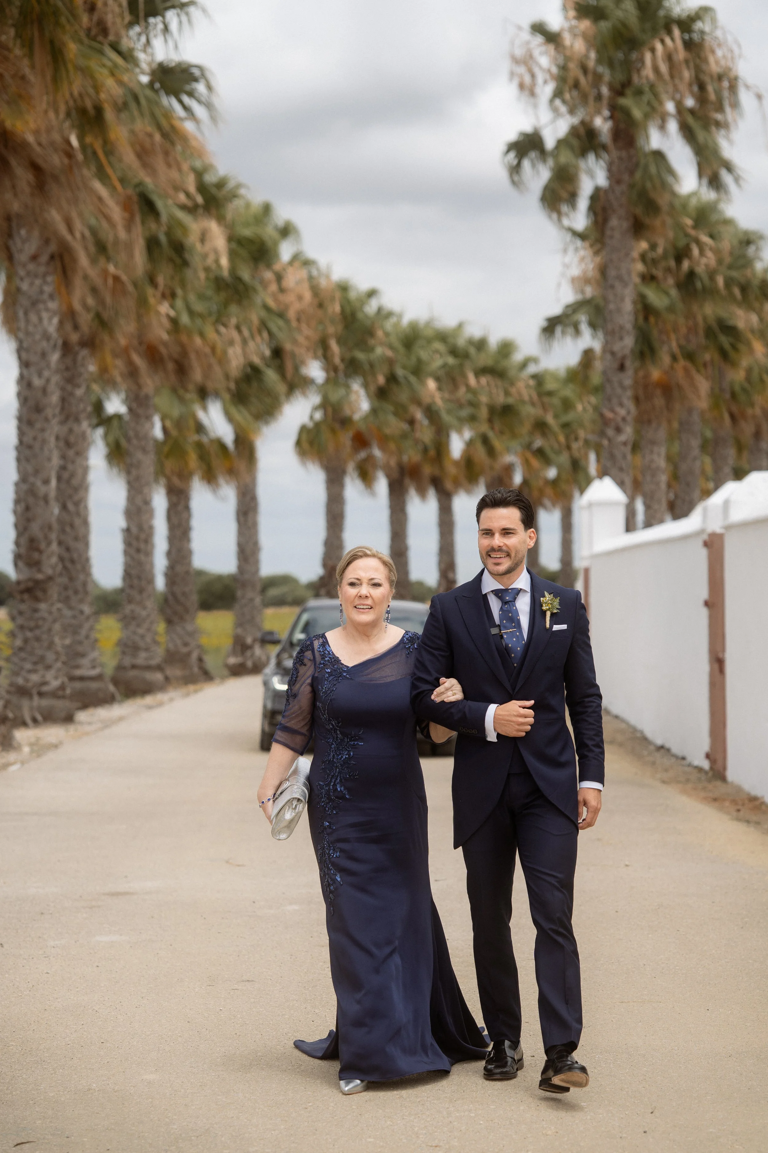 Una mujer mayor y un hombre joven en un camino con palmeras, probablemente en una boda, ambos vestidas formalmente y sonriendo.