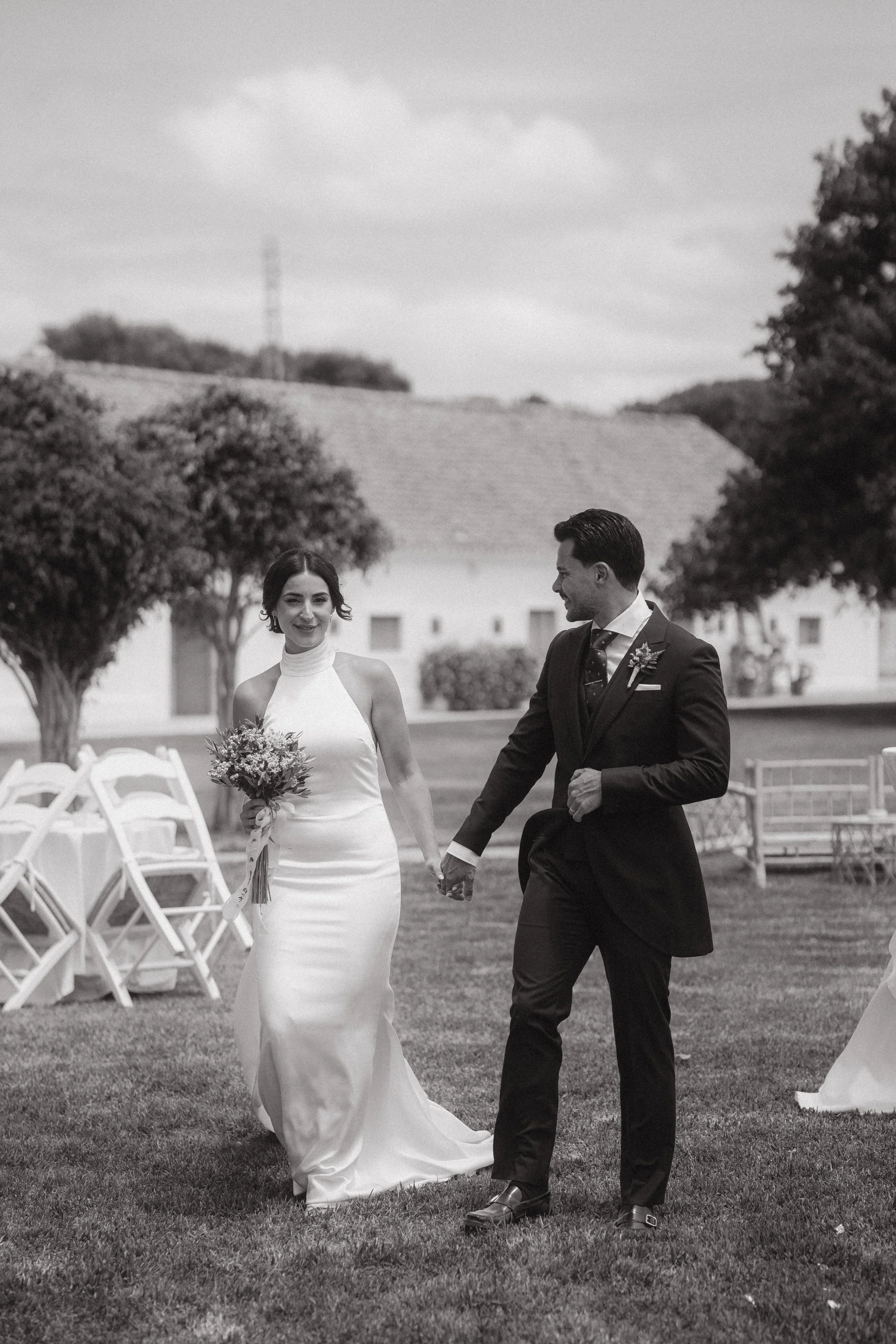Pareja de novios caminando juntos en una boda al aire libre en un campo, con árboles y una casa en el fondo, en blanco y negro.