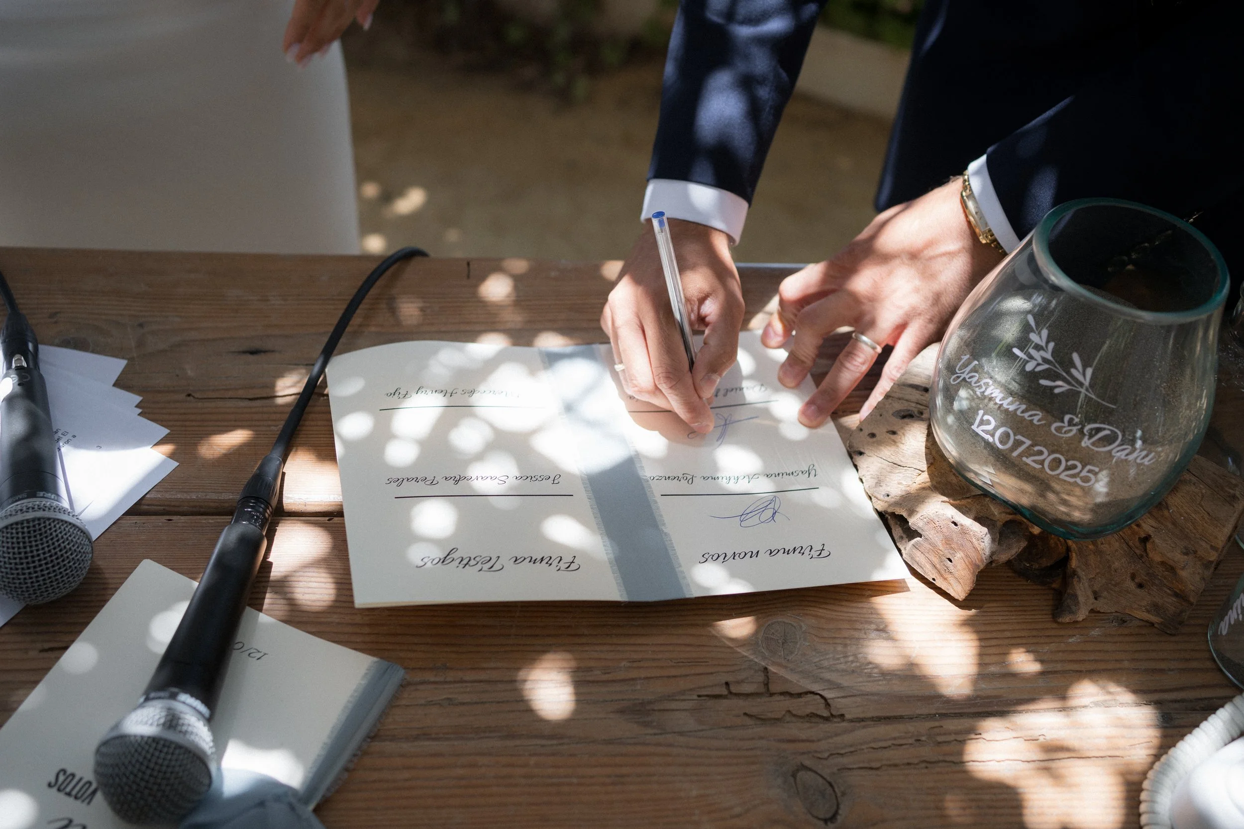 Una pareja firma un documento de boda al aire libre, rodeados de sombra y luz natural, con un vaso decorativo que tiene nombres y una fecha