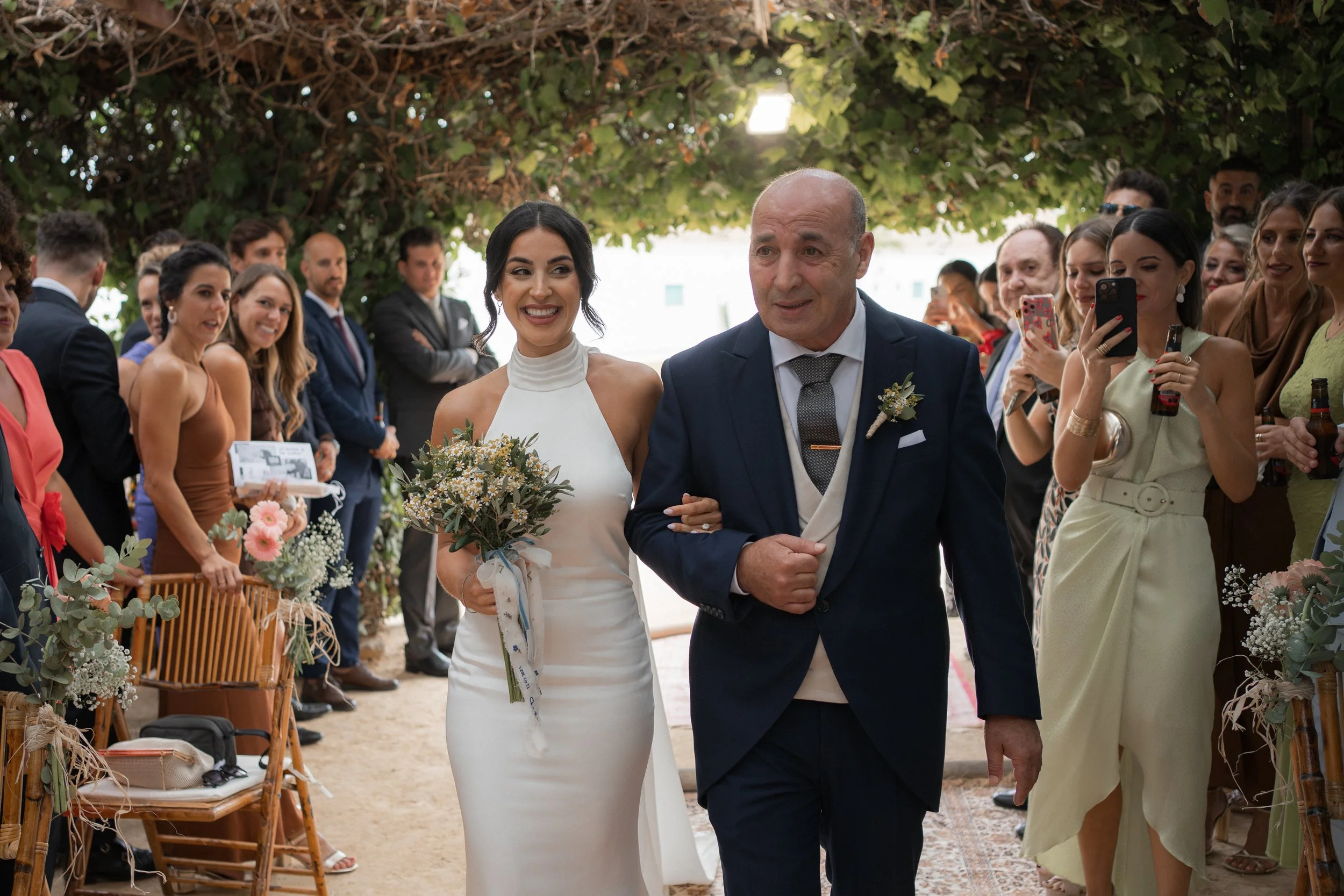 Una boda al aire libre con la novia sonriendo y acompañada por un hombre mayor mientras caminan por el pasillo. La ceremonia está rodeada de amigos y familiares que toman fotos y observan con alegría.