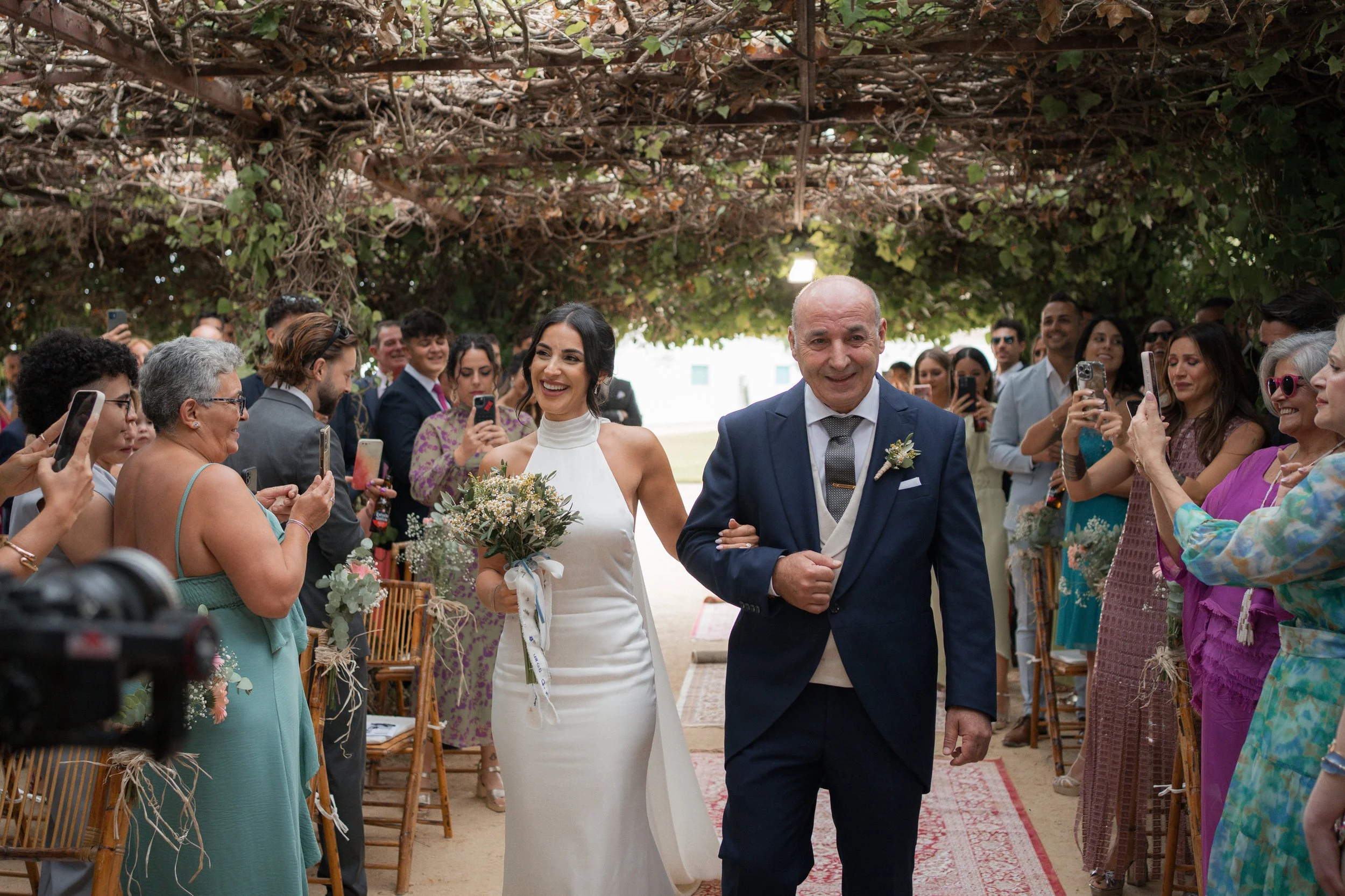 Pareja de novios, una mujer con vestido blanco y una mujer con saco y corbata, en una ceremonia de boda, rodeados de familiares y amigos que toman fotos.