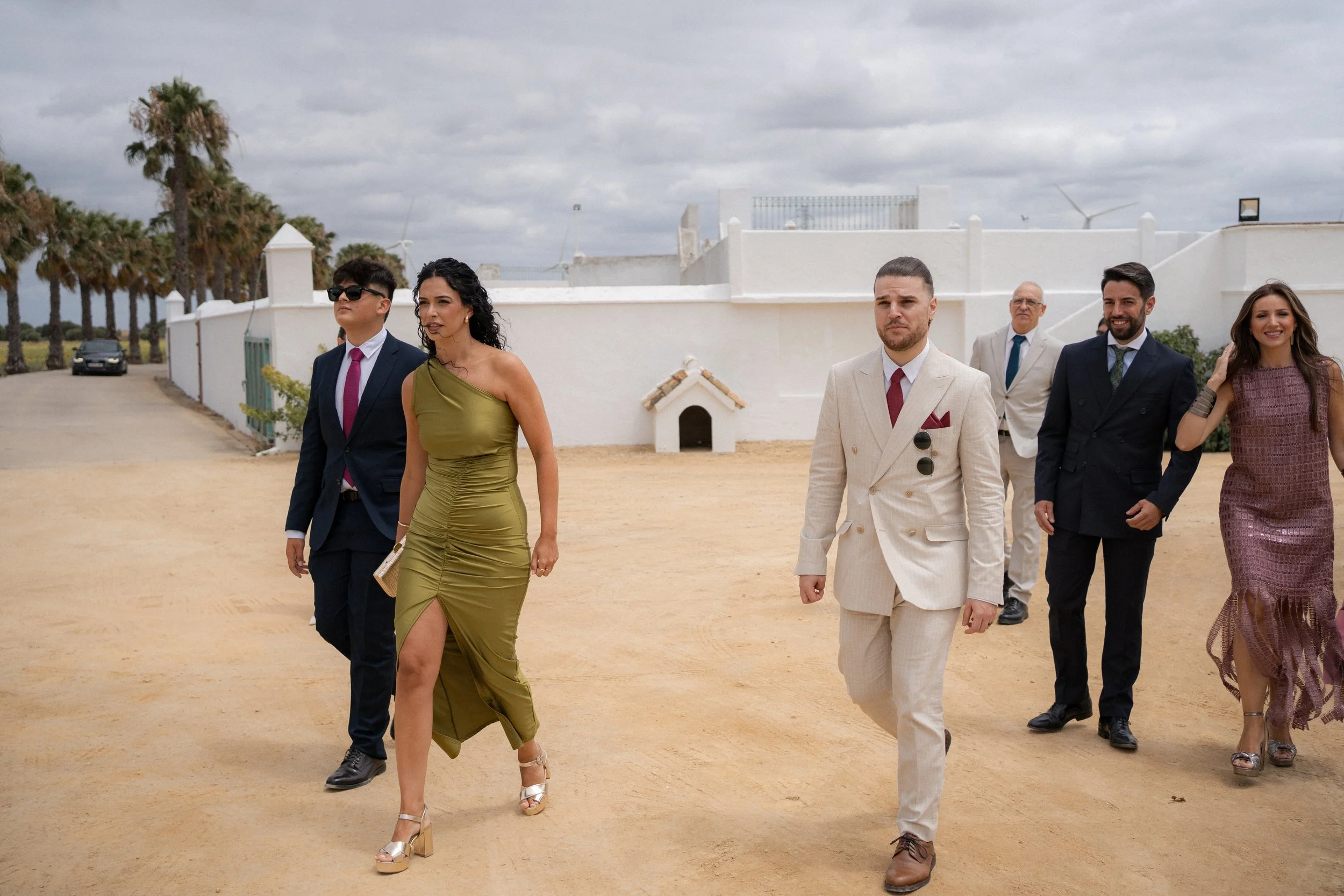 Grupo de seis personas vestidas formalmente caminando en un camino de tierra con palmeras al fondo y un cielo nublado.