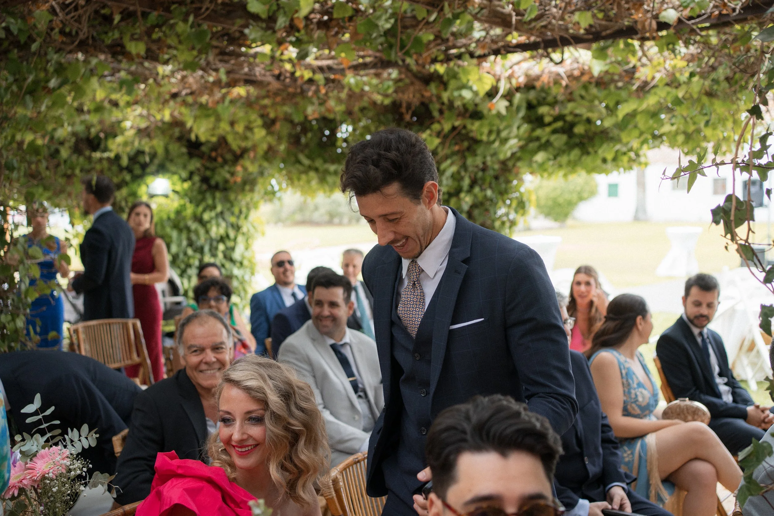 Celebración al aire libre con personas vestidas formalmente, en un lugar decorado con plantas y flores, participando en lo que parece ser un evento especial como una boda o ceremonia.