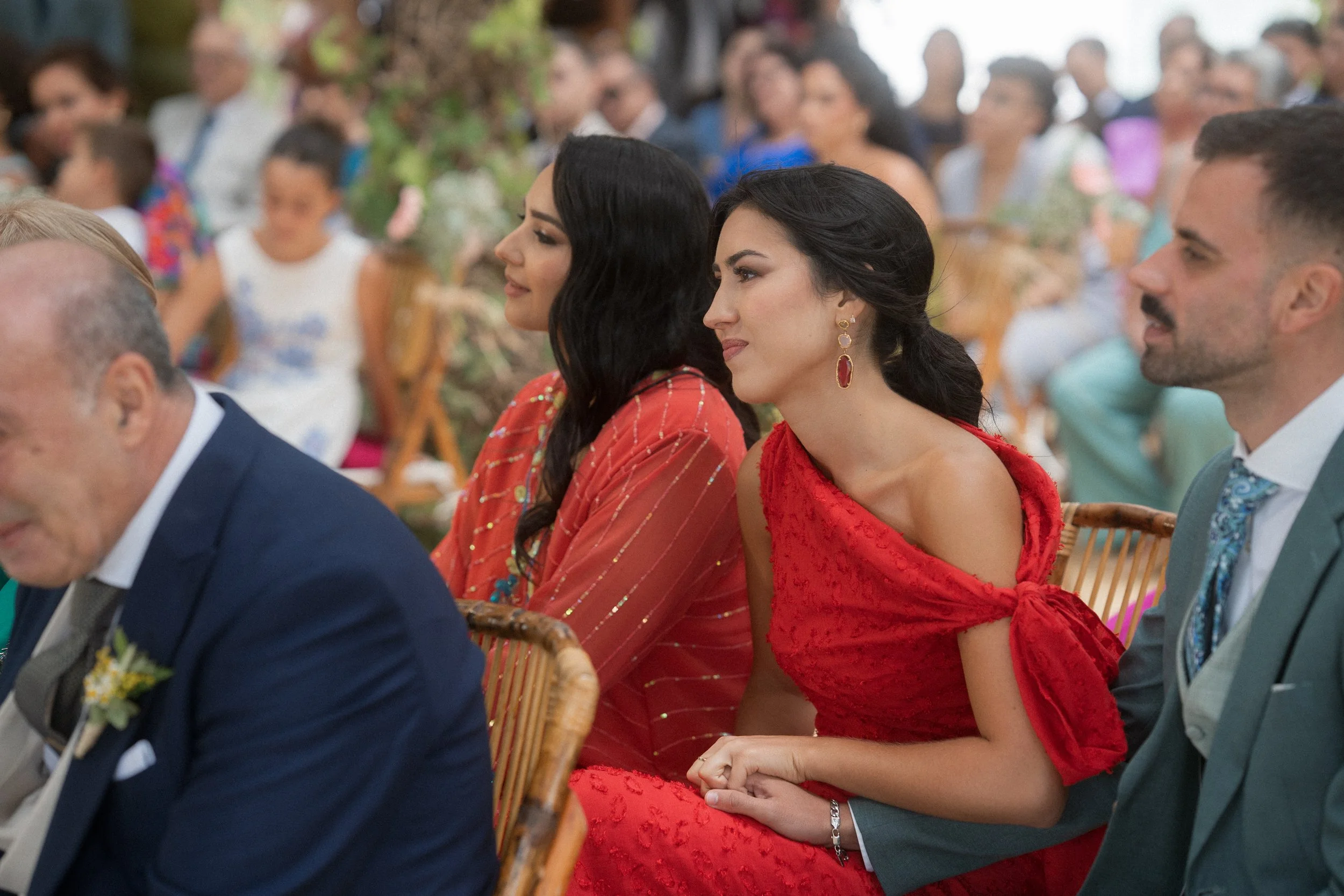 Personas sentadas en una ceremonia, algunas con vestidos elegantes, en un entorno decorado con flores.
