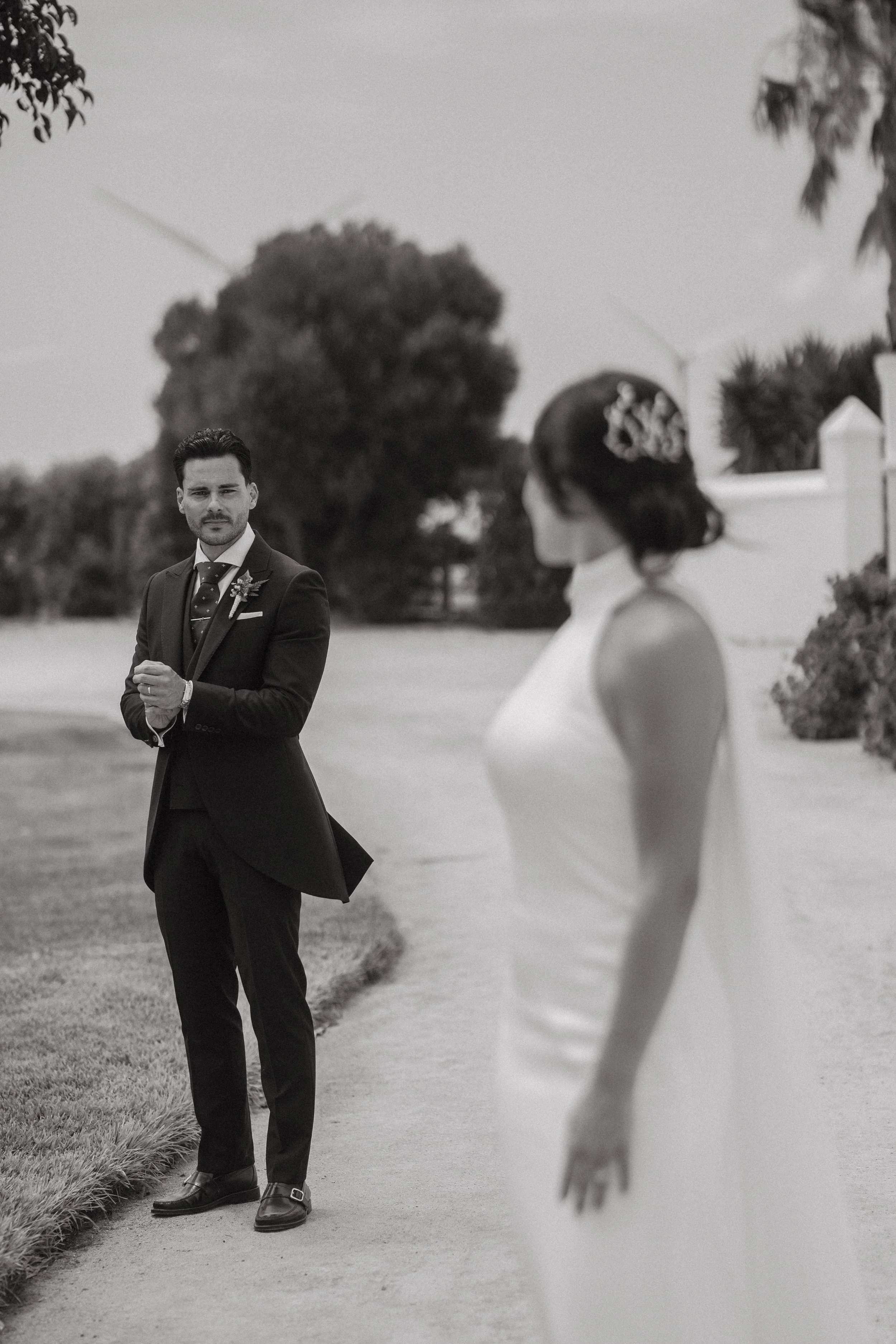 Un hombre vestido con traje negro observa a una mujer en un vestido blanco, en un ambiente al aire libre con árboles y una cerca de fondo.