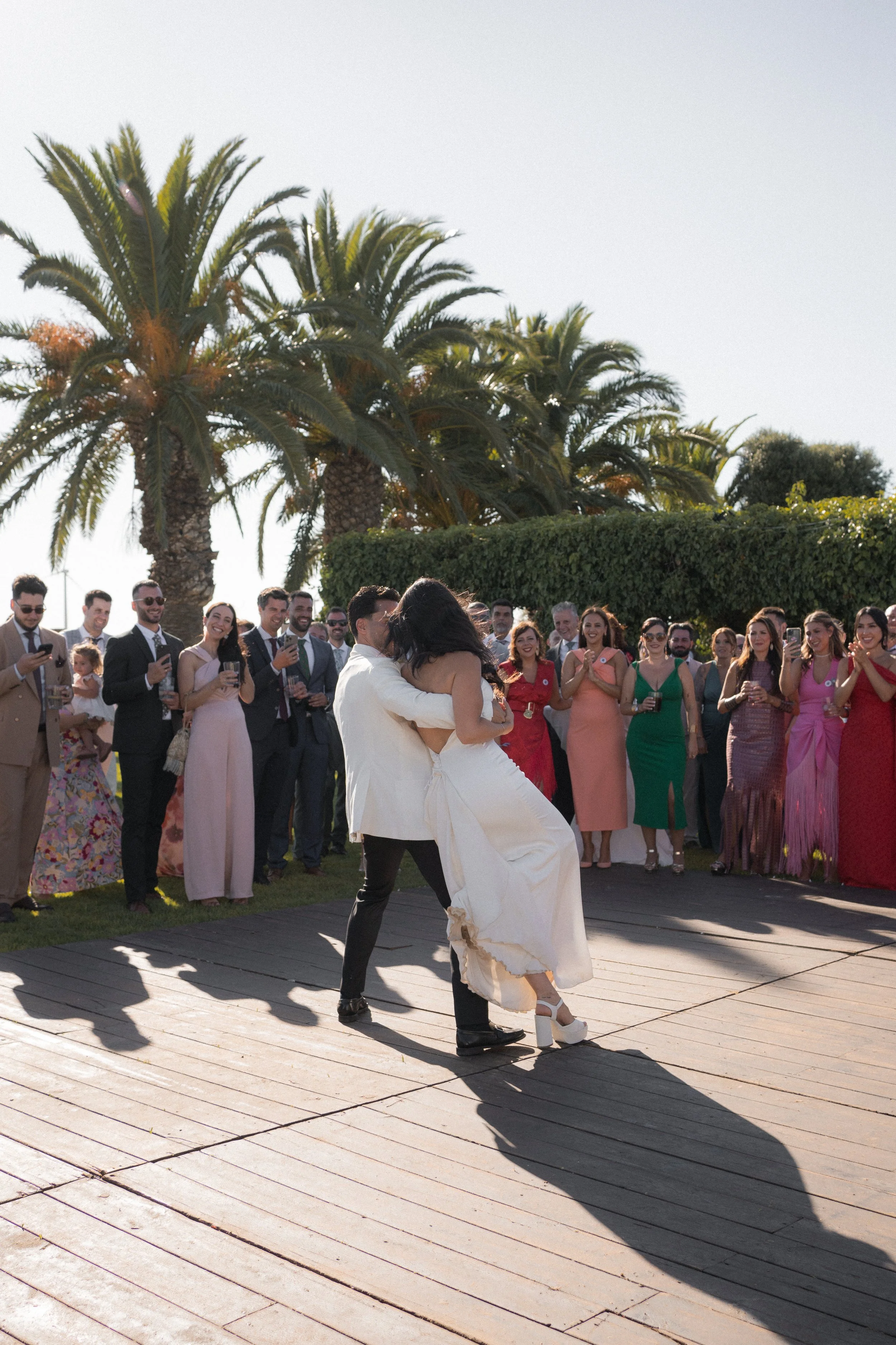 Pareja bailando en una boda al aire libre, rodeados de invitados con palmeras en el fondo.