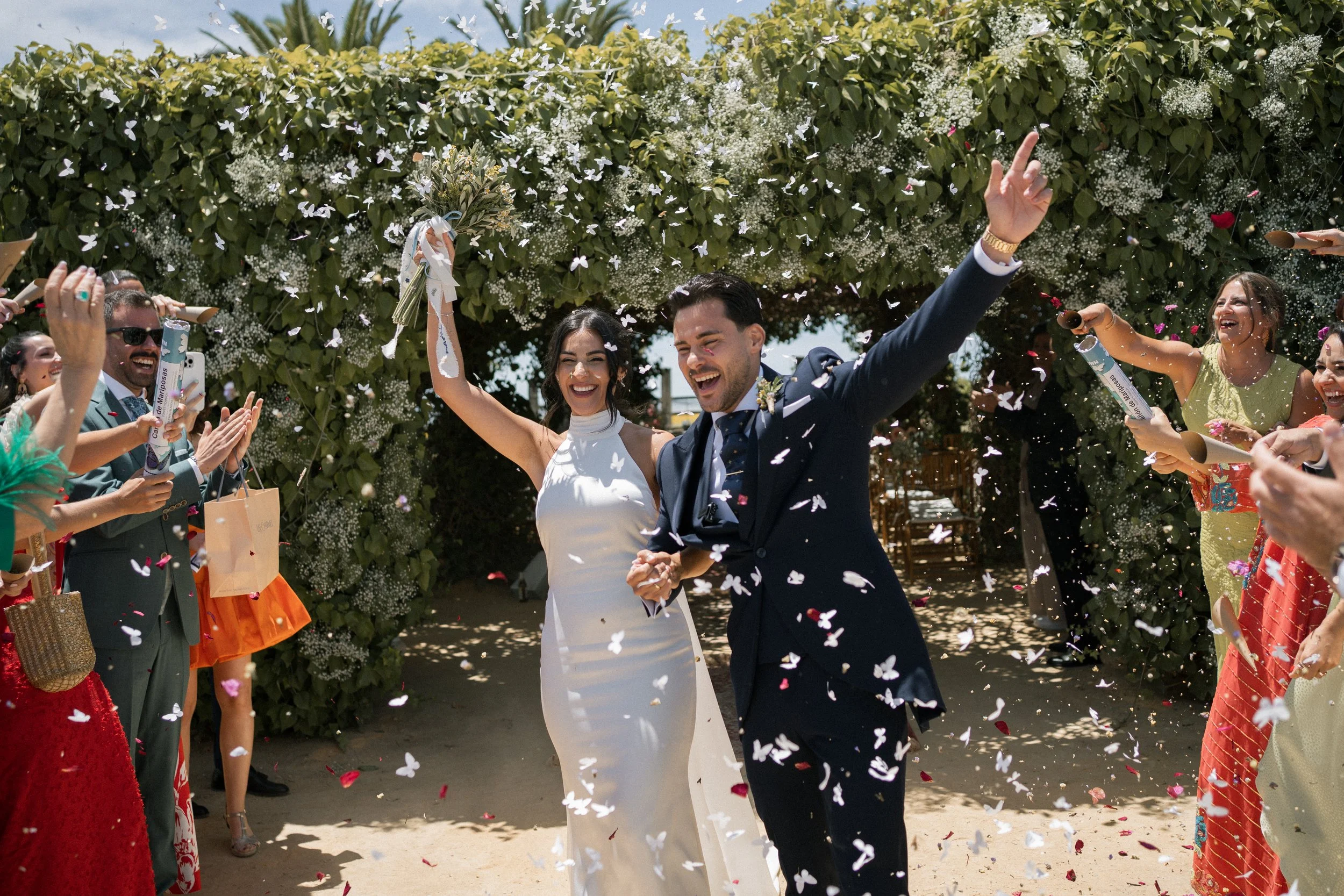 Pareja de recién casados celebrando su boda bajo un arco de flores, rodeados de amigos lanzando confeti.