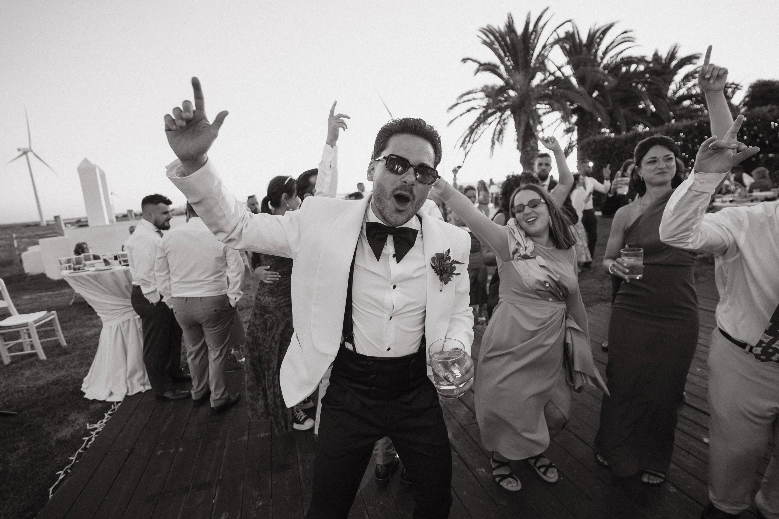 Personas bailando y disfrutando en una fiesta al aire libre, vestido formal, en un día soleado con palmeras y molinos de viento en el fondo.