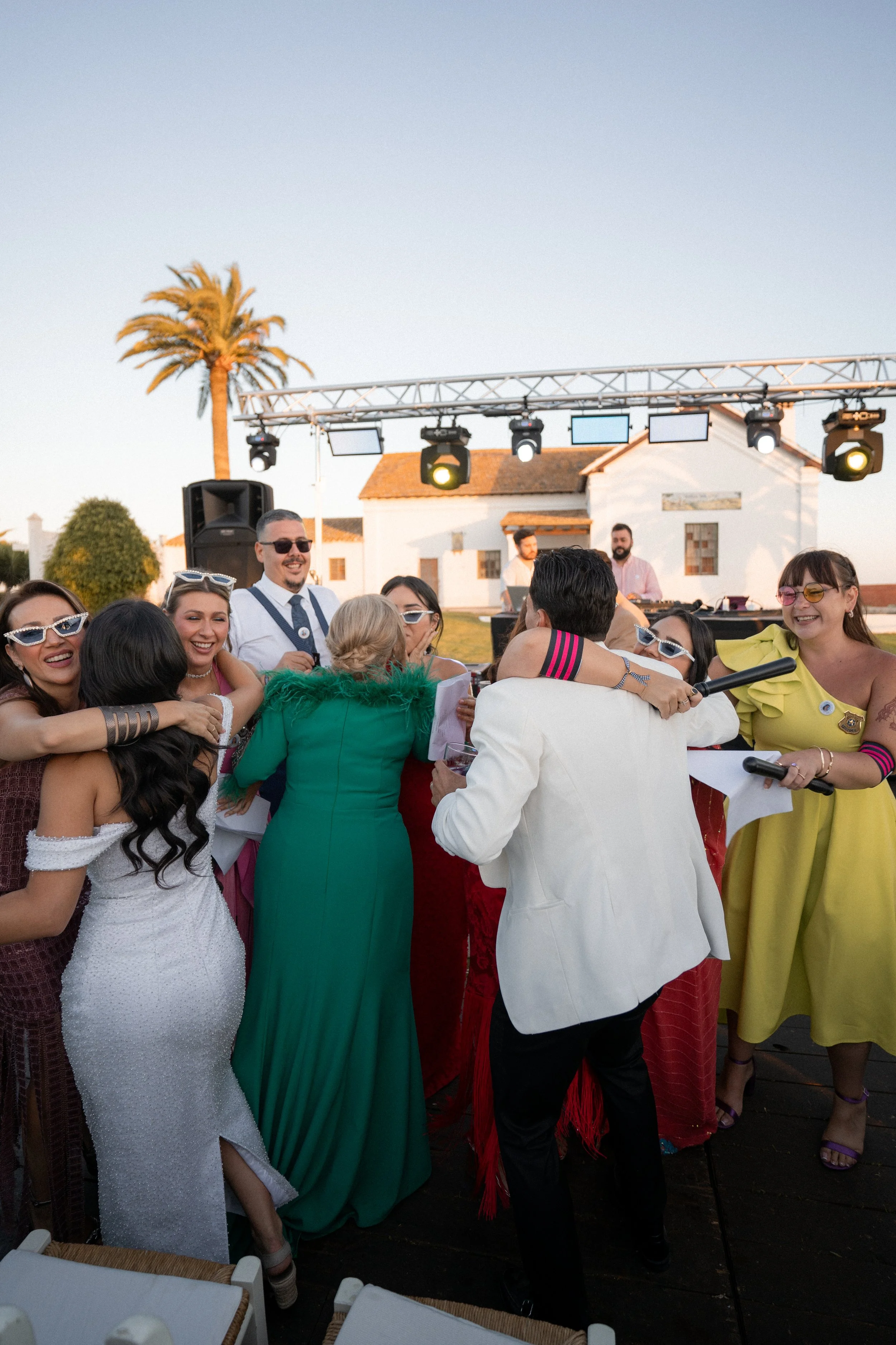 Un grupo de personas en una fiesta abrazándose y sonriendo, con un DJ en el escenario de fondo y luces en un entorno al aire libre.