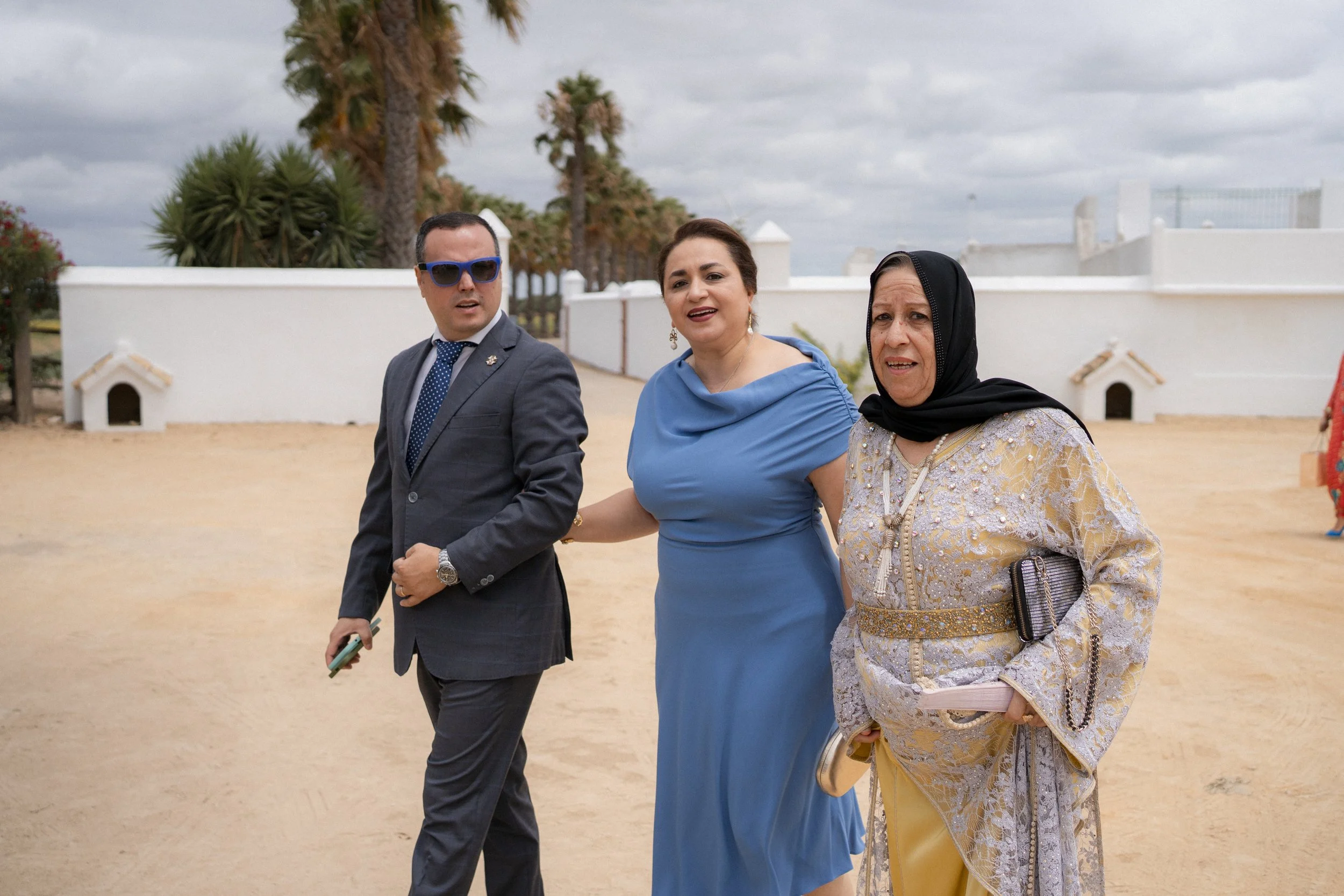 Tres personas caminando al aire libre, un hombre con traje y gafas de sol, una mujer en vestido azul y otra mujer en vestido claro con pañuelo en la cabeza. Detrás hay árboles y un muro blanco, el cielo está nublado.