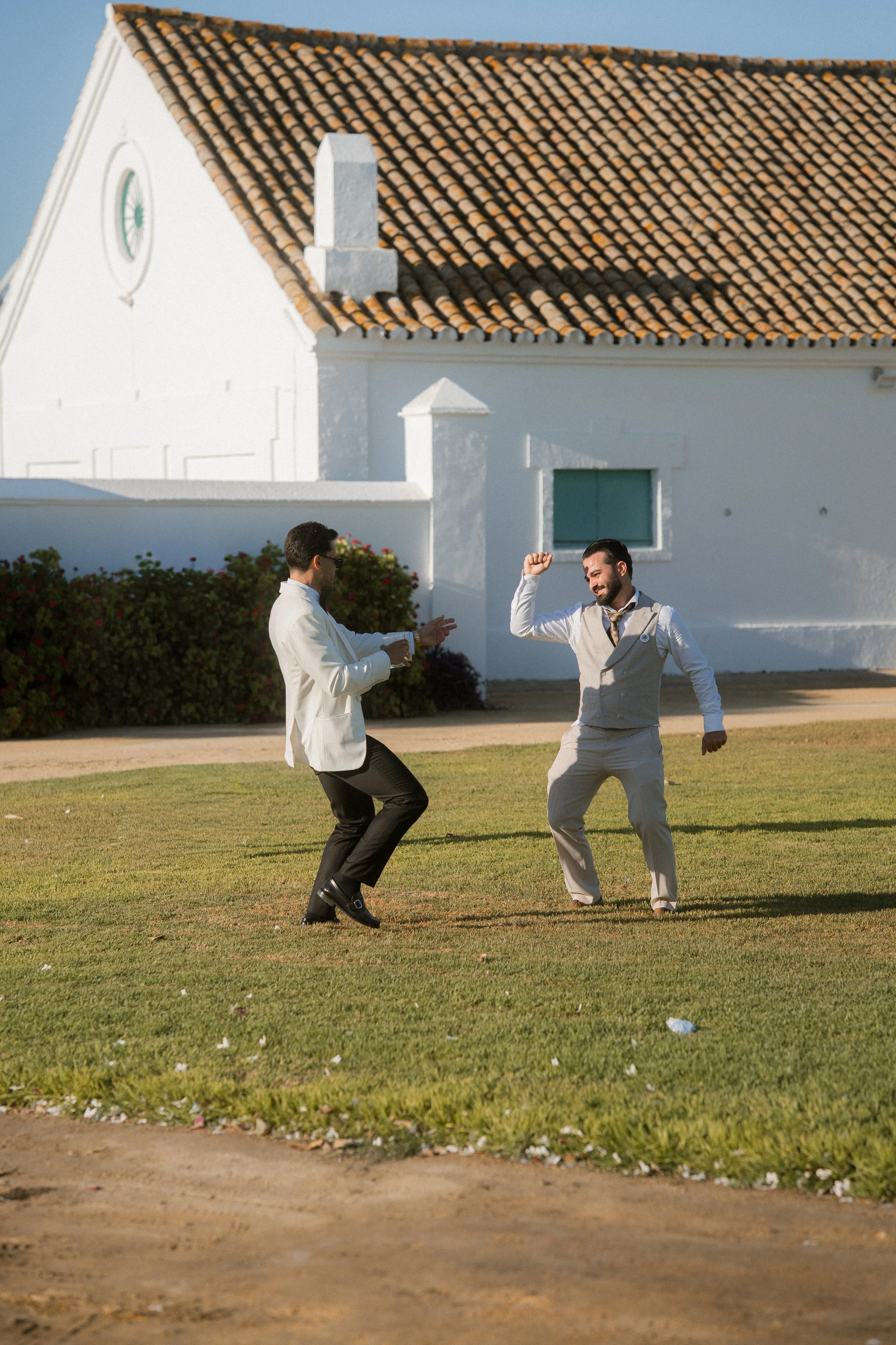 Dos hombres bailando y divirtiéndose en un césped frente a una iglesia blanca con techo de tejas.