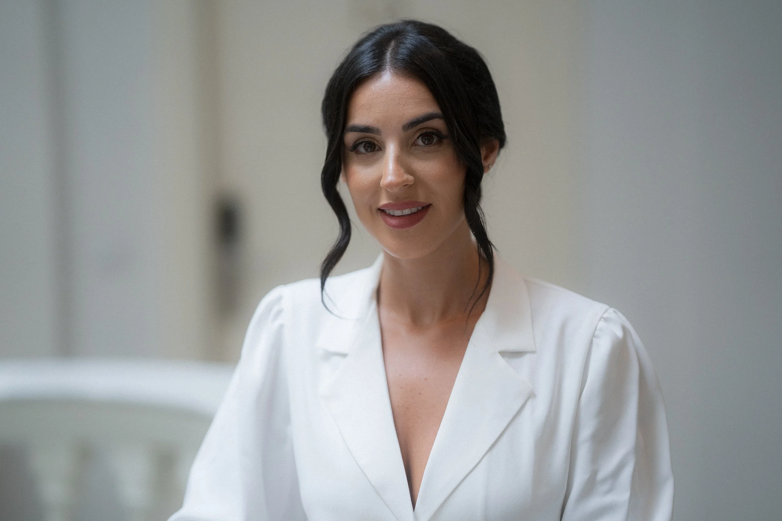 Mujer con cabello oscuro y rizado, sonriendo, vestida con bata blanca, en un entorno clínico o hospitalario.