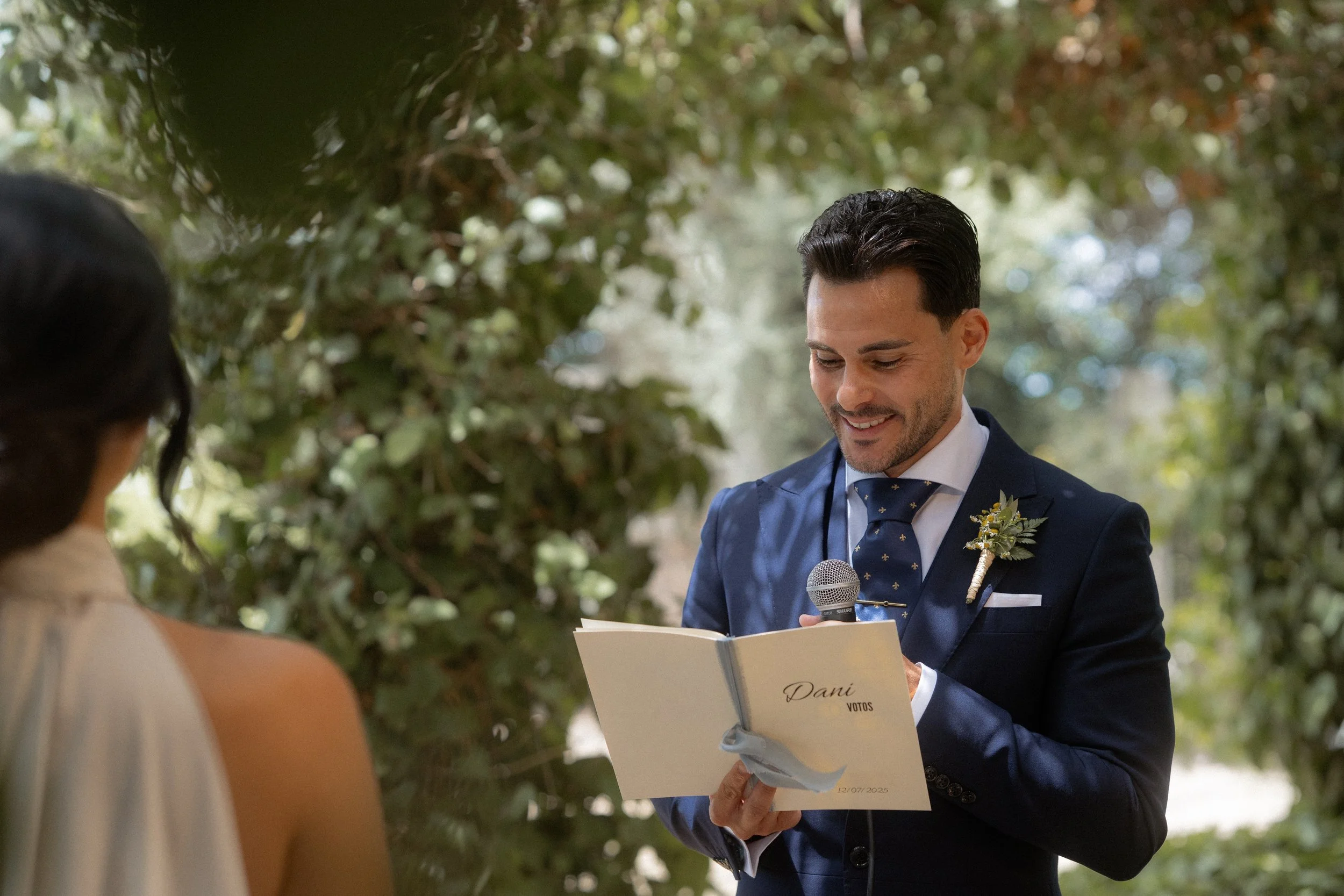 Hombre en traje de boda leyendo votos en una ceremonia al aire libre, con una mujer frente a él, en un entorno natural con árboles.