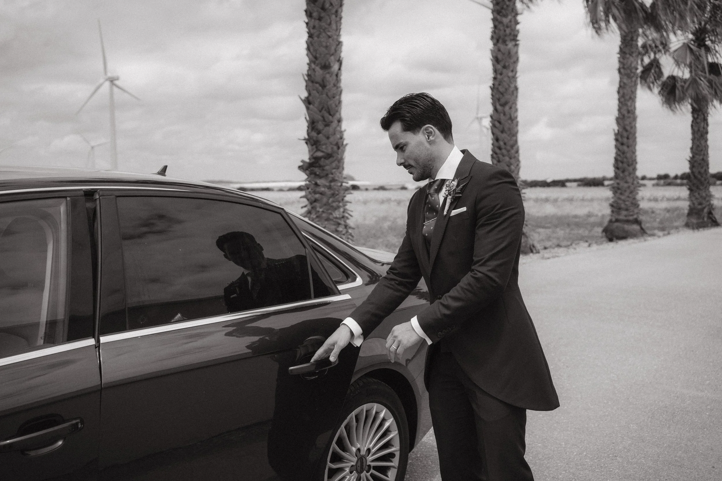 Un hombre vestido con traje elegante se está acercando a un coche elegante, en un entorno con palmeras y molinos de viento, en un día nublado.