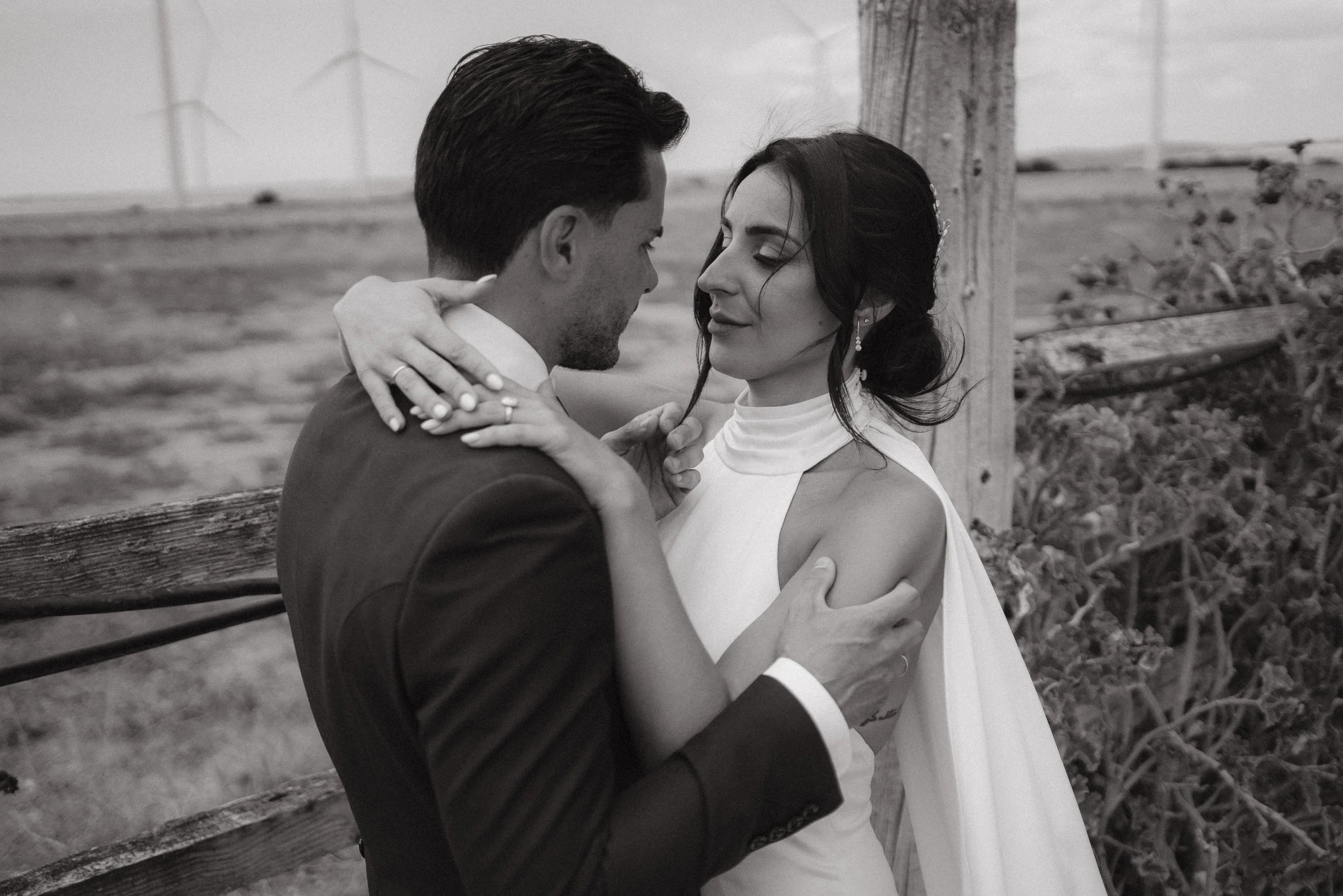 Una pareja de esposos se abraza frente a una cerca de madera en un campo abierto, en un momento romántico.