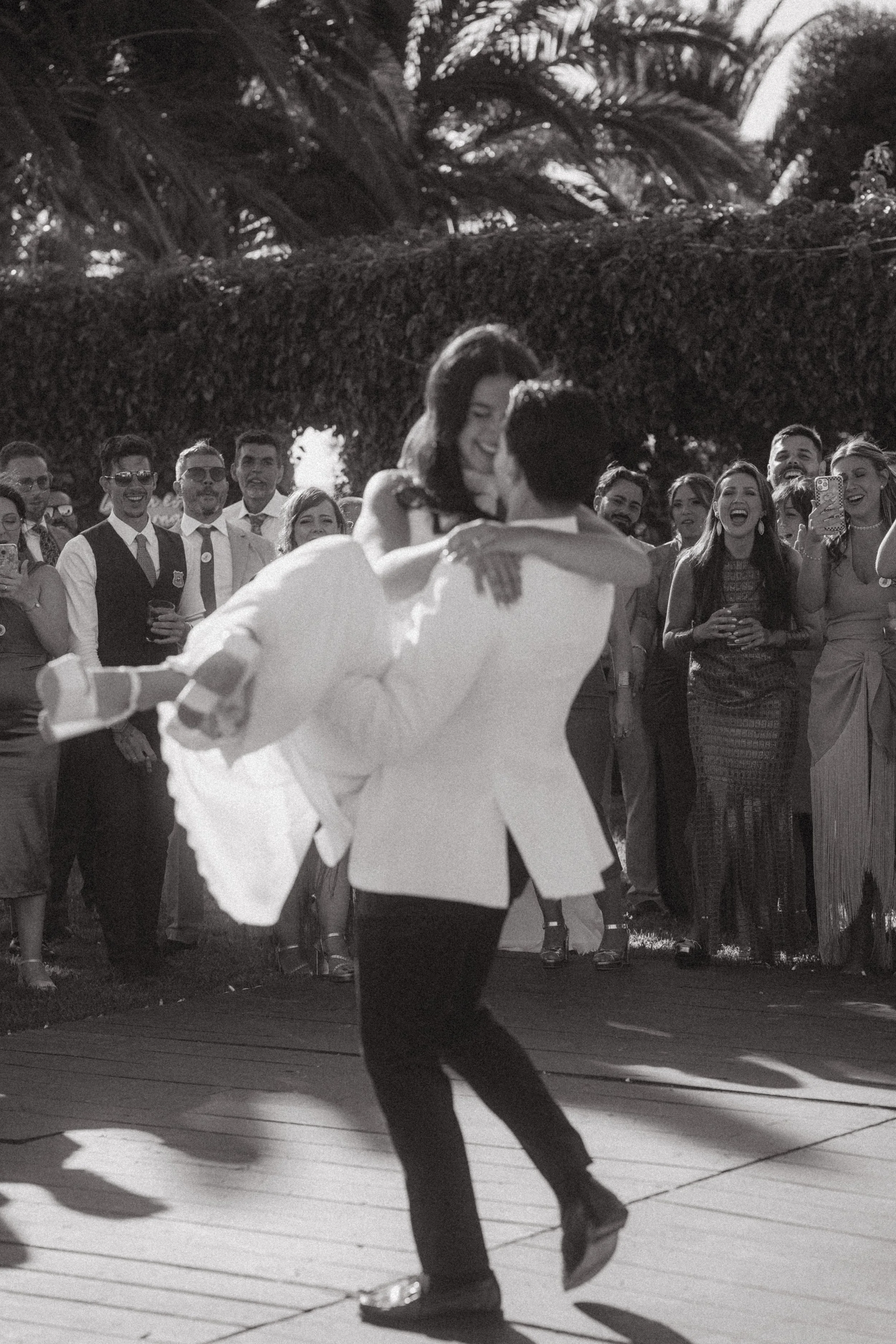 Pareja bailando en una celebración mientras un grupo de amigos los observa y celebra, en un ambiente al aire libre con vegetación en el fondo.