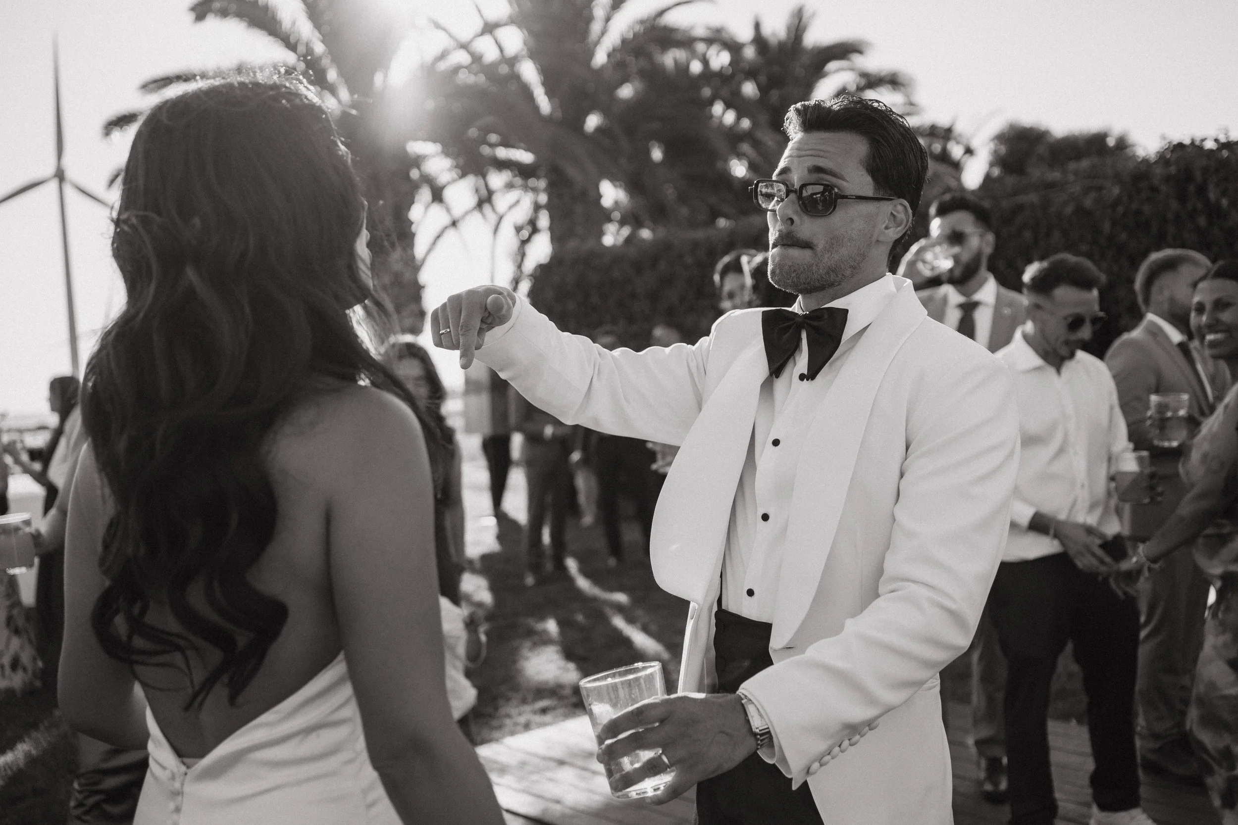 Hombre con lentes y traje blanco con pajarita, sosteniendo un vaso, bailando en una fiesta al aire libre con un grupo de personas, palmeras y viento de fondo.
