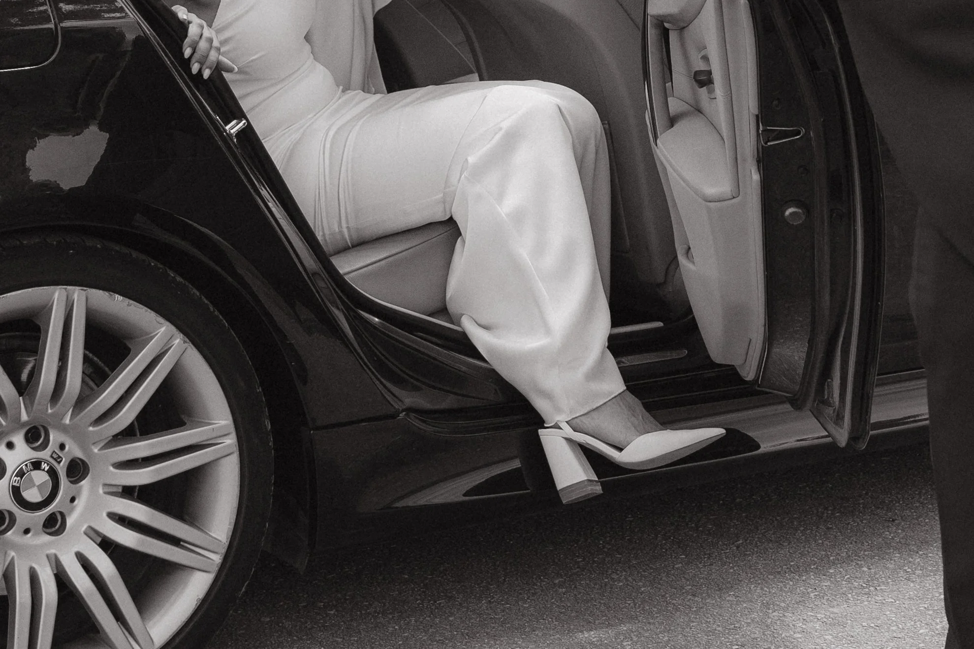 Mujer vestida de blanco con tacón alto en el asiento de un coche negro, con la puerta abierta.