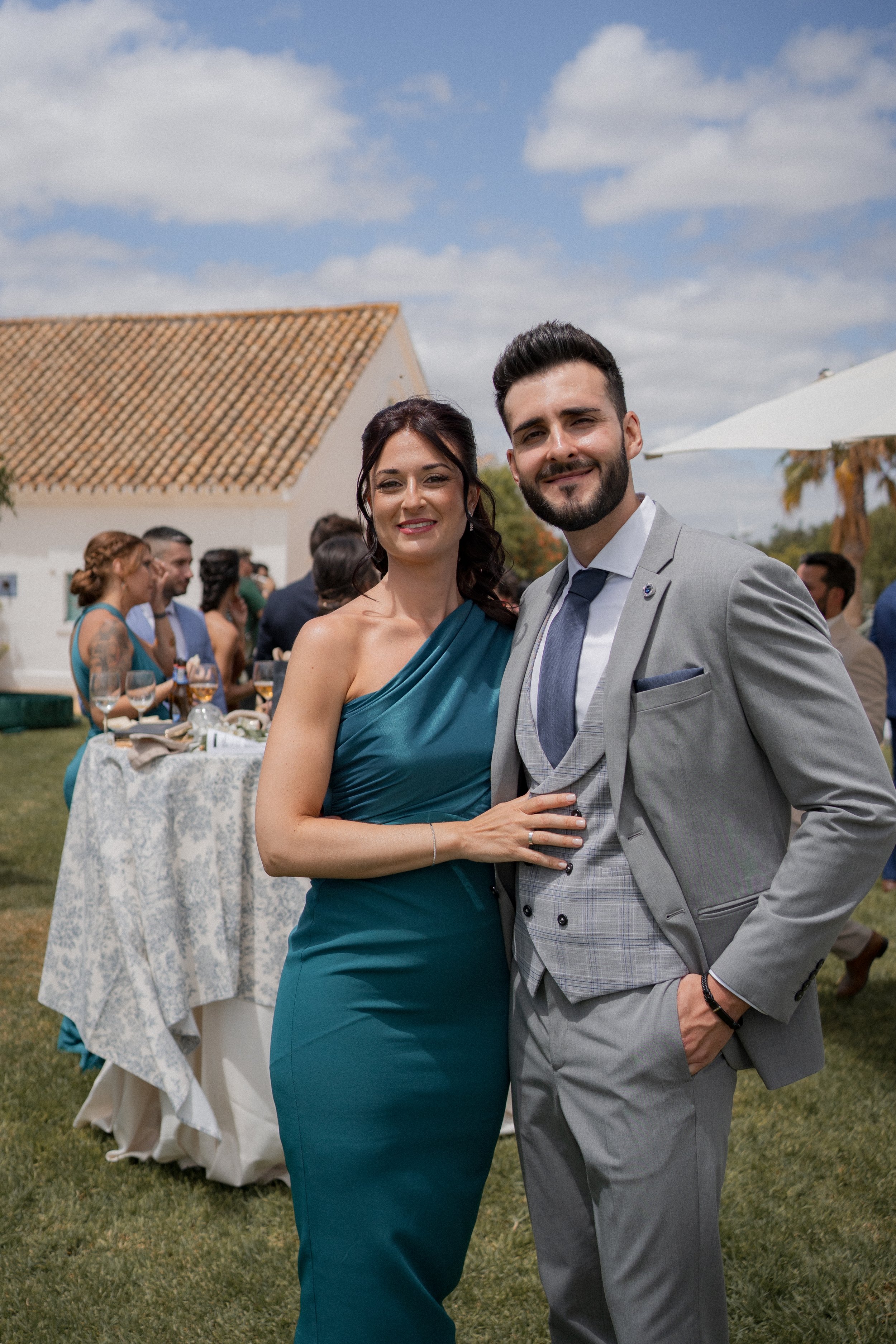 Pareja sonriente en celebración al aire libre, con otros invitados y mesas decoradas en el fondo.