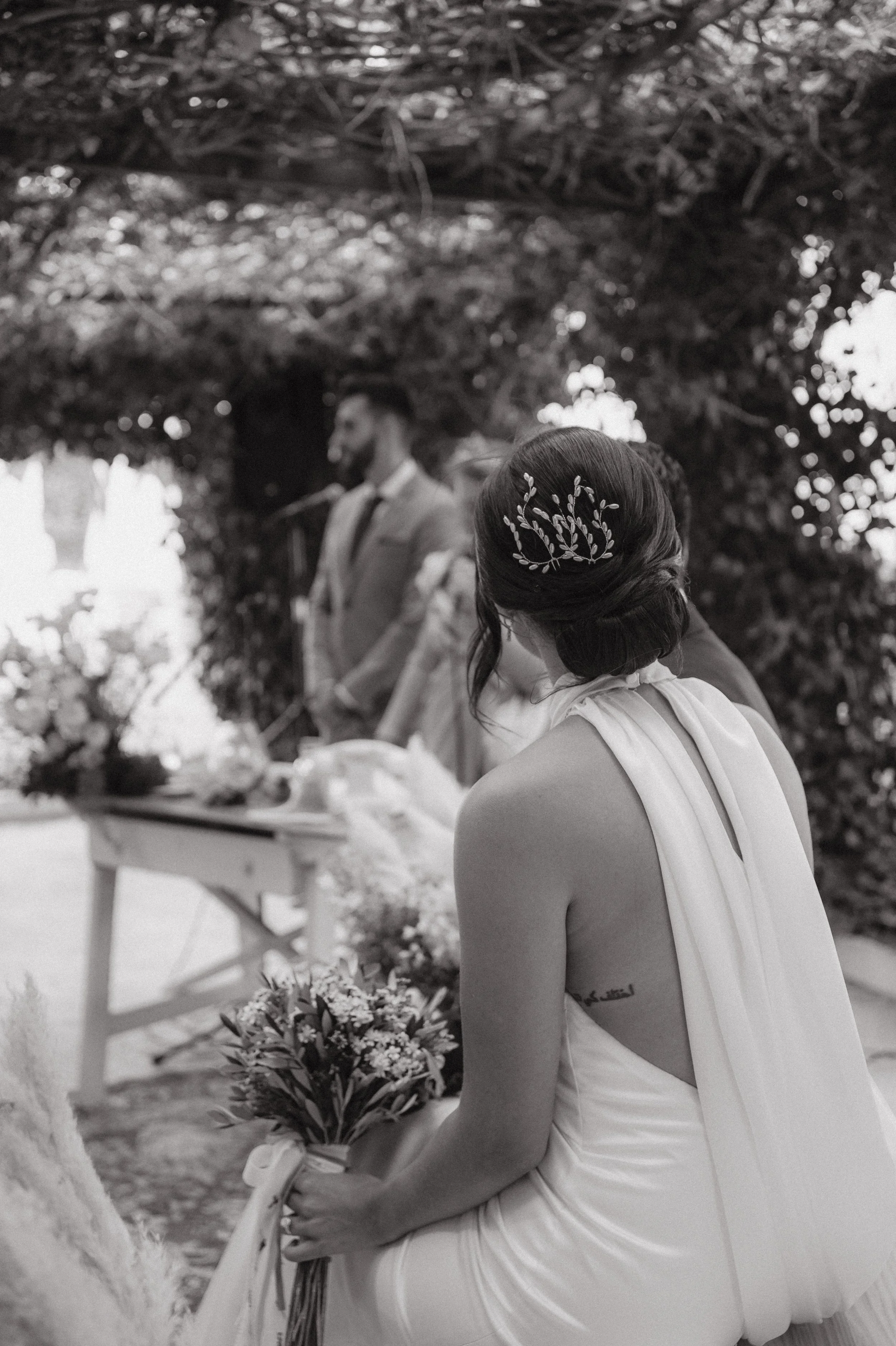 Mujer en vestido de novia con cabello recogido y adorno de ramas, sosteniendo un ramo de flores, en una ceremonia de boda al aire libre, con un hombre en el fondo y un altar decorado con flores.