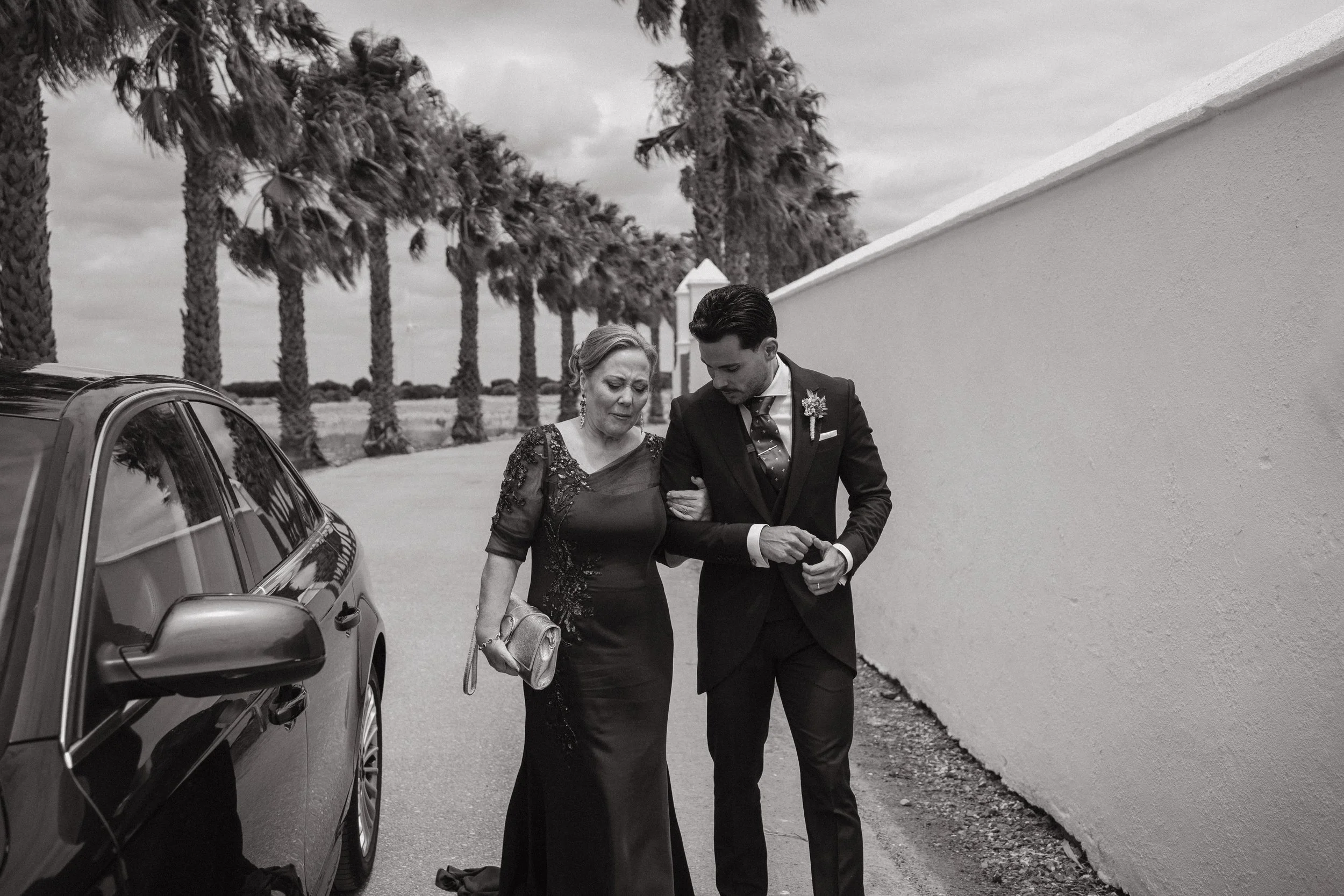 Un hombre joven ayuda a una mujer mayor a caminar por la calle, al lado de un coche y con palmeras en el fondo, en un entorno al aire libre.