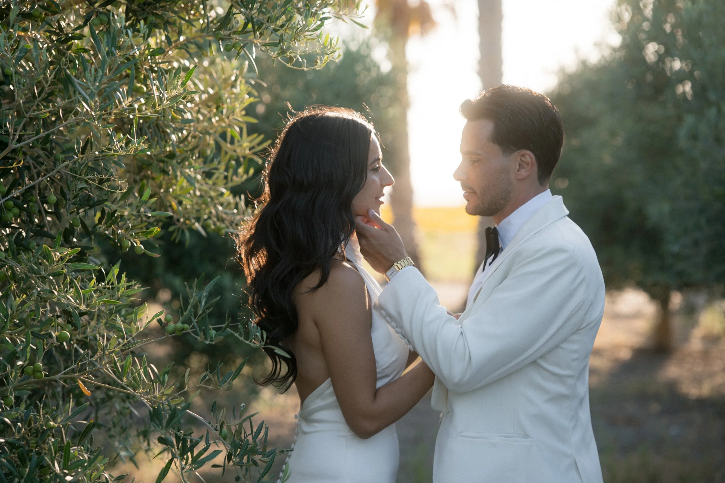 Una pareja en una ceremonia romántica al aire libre durante el atardecer, con árboles verdes en el fondo, ella con vestido blanco y él con saco blanco y pajarita negra, ella mira a él mientras él la toca suavemente.