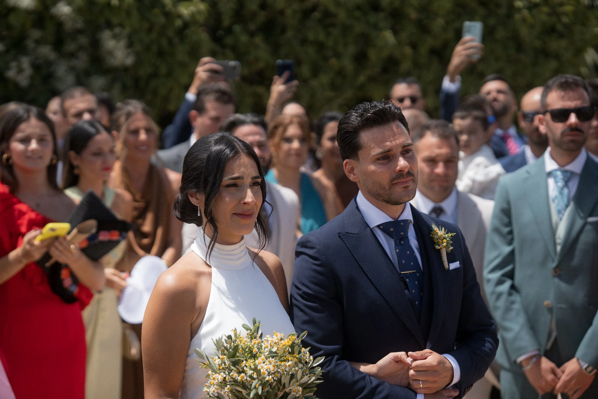 Pareja en una boda, la mujer sostiene un ramo de flores y el hombre viste un traje azul oscuro, con fondo de invitados