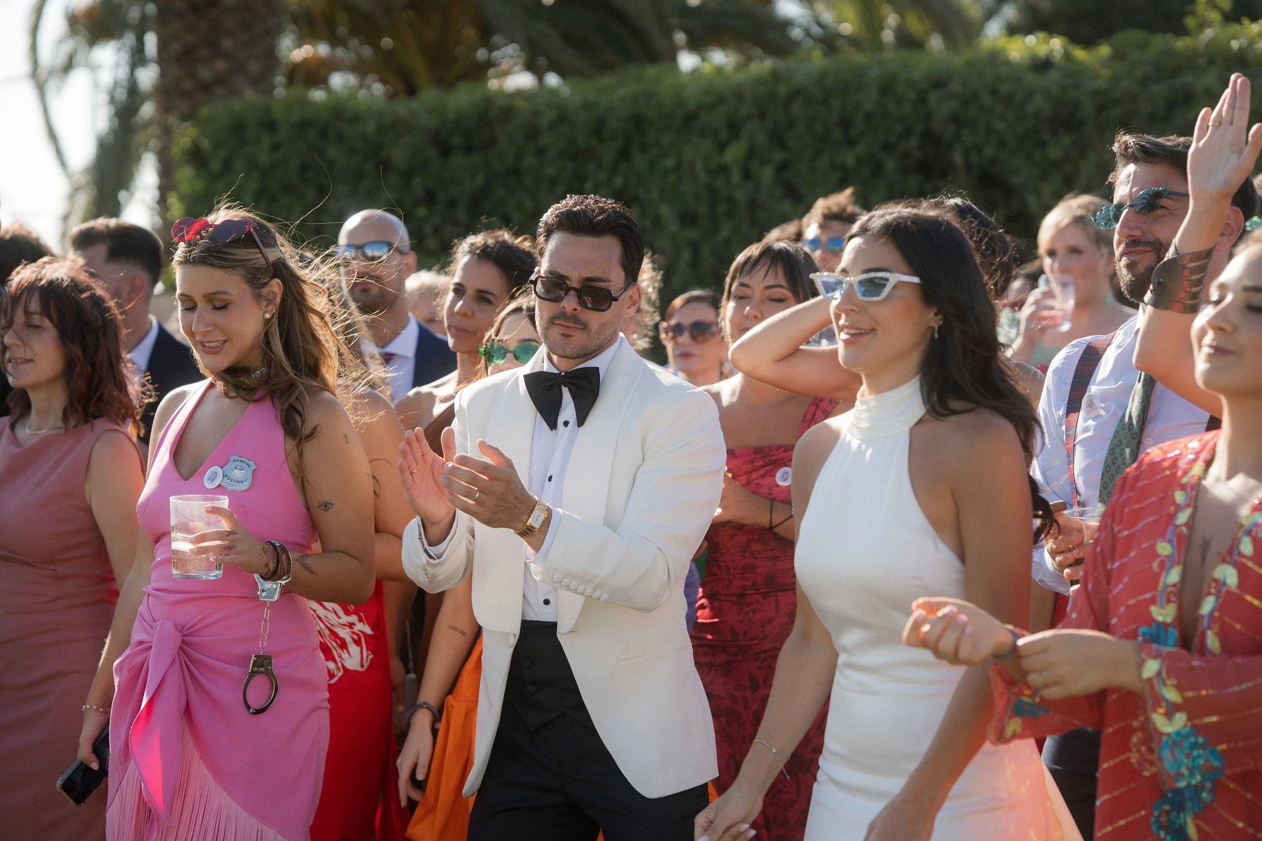 Grupo de personas en un evento al aire libre, algunas con gafas y ropa elegante, parecen disfrutar y celebrar.