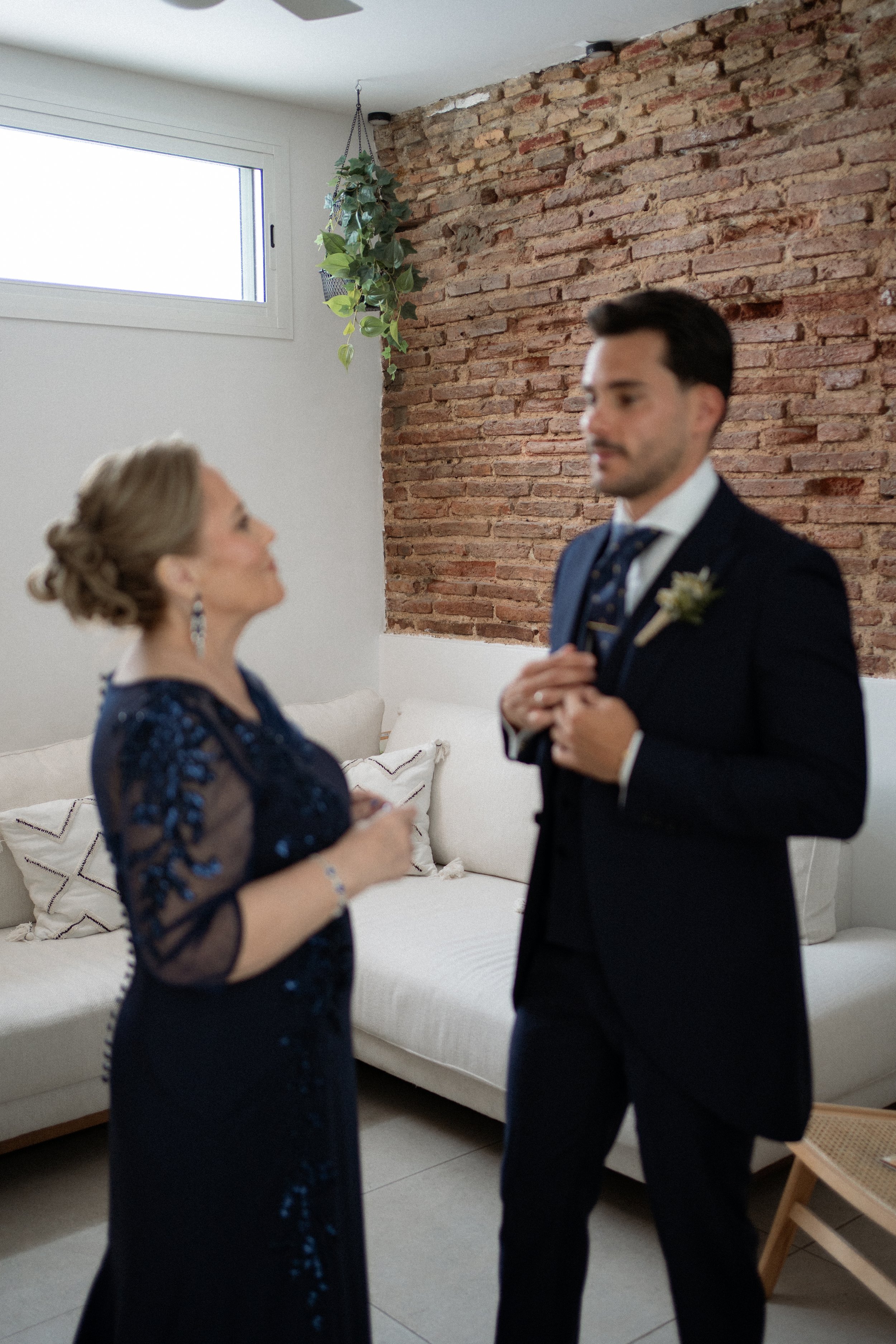Una mujer y un hombre vestidos formalmente en una sala con pared de ladrillos expuestos y sofá claro, parecen estar en una conversación seria o emotiva.