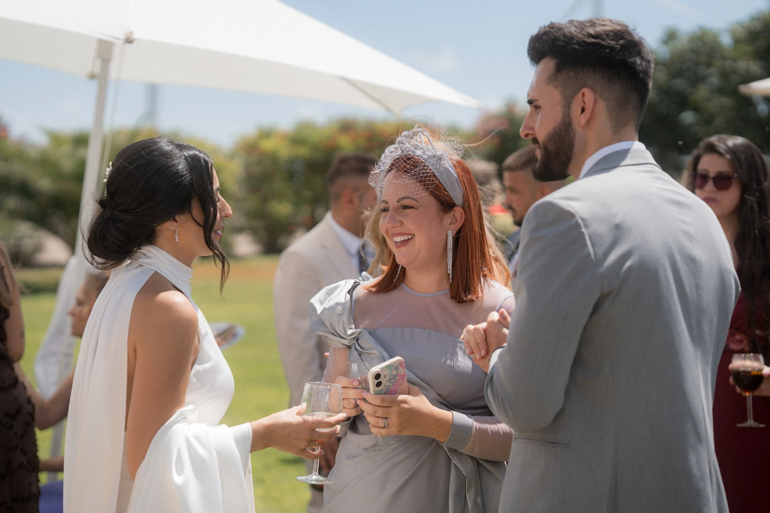 Grupo de personas en una recepción de boda al aire libre, con parejas y amigos disfrutando y socializando, ambiente soleado con paraguas y árboles en el fondo.