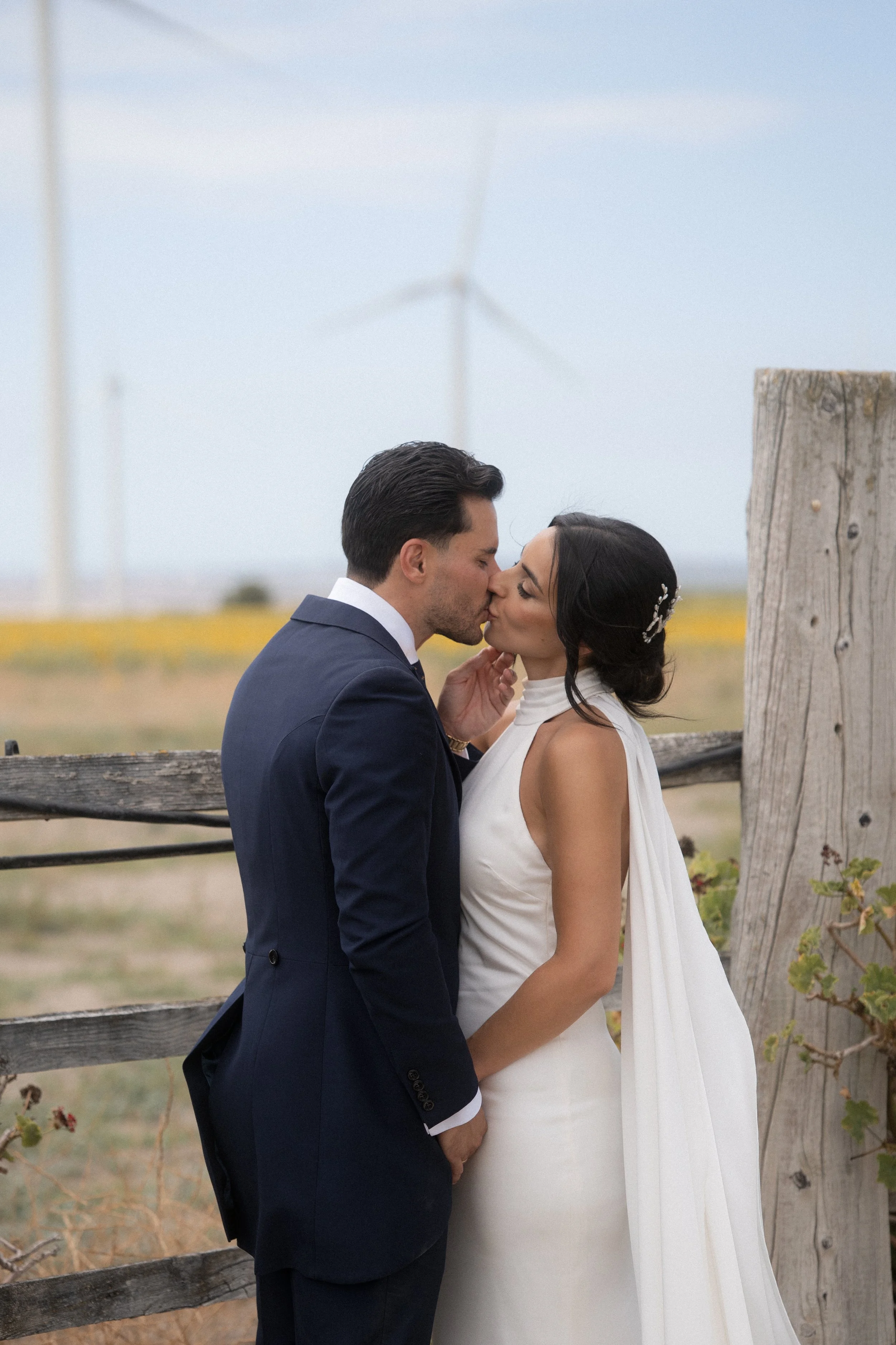 Pareja de recién casados besándose en un campo con molinos de viento al fondo.
