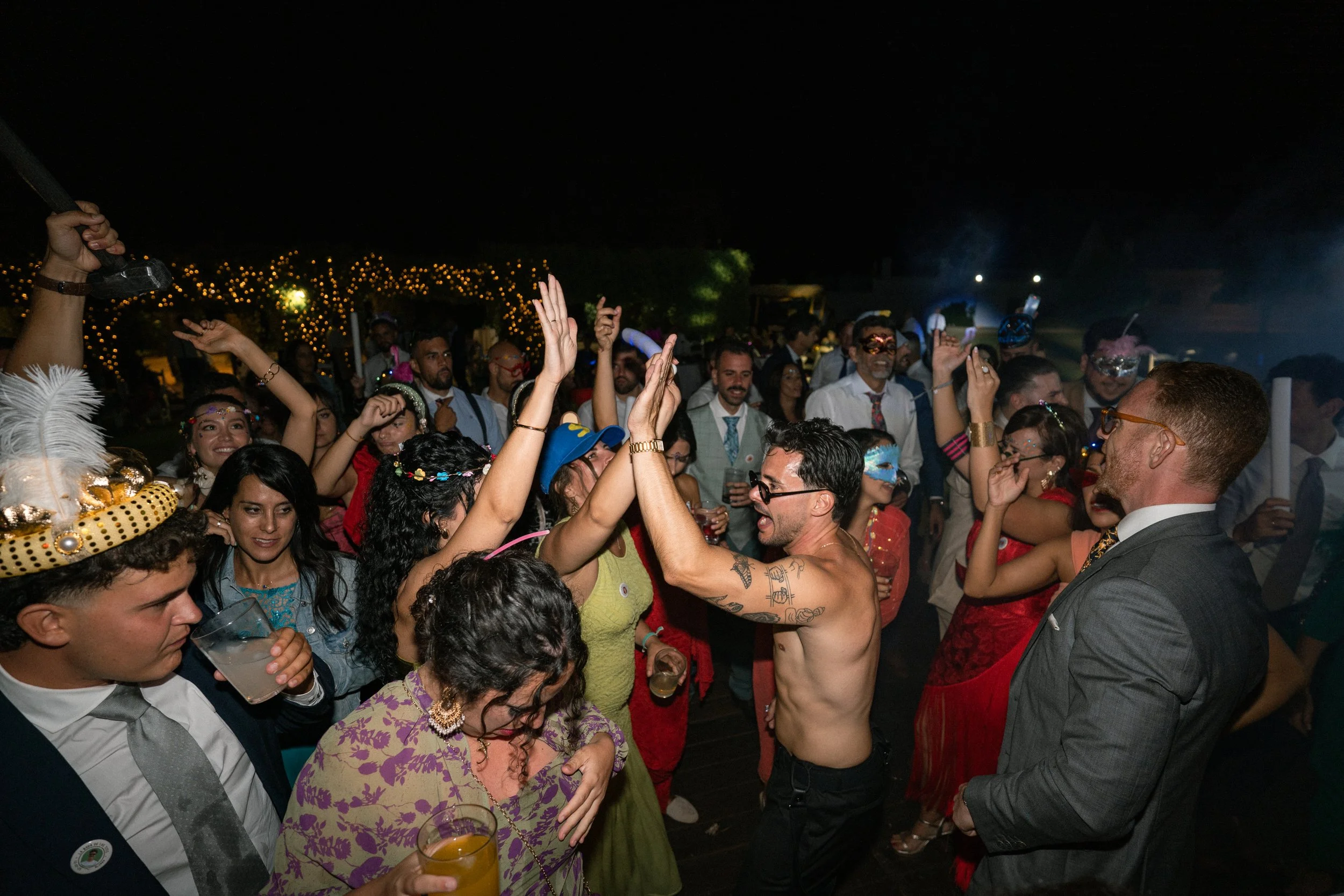 Grupo de personas bailando y divirtiéndose en una boda nocturna, algunos llevan disfraces y máscaras, disfrutando de la música y la compañía.