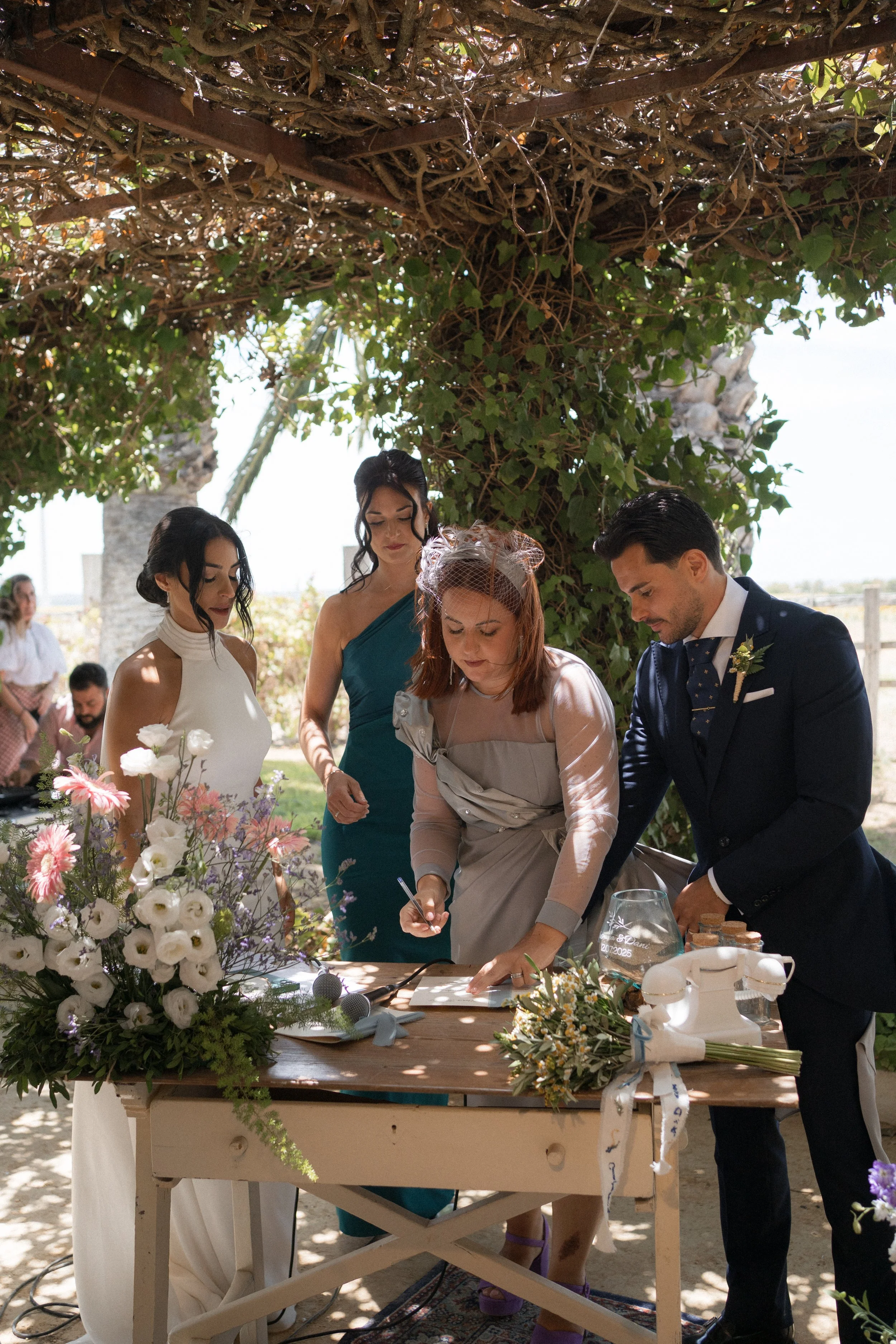 Una ceremonia de matrimonio al aire libre con una pareja y dos mujeres, decoraciones florales y una mesa con elementos para la ceremonia bajo un árbol con follaje.