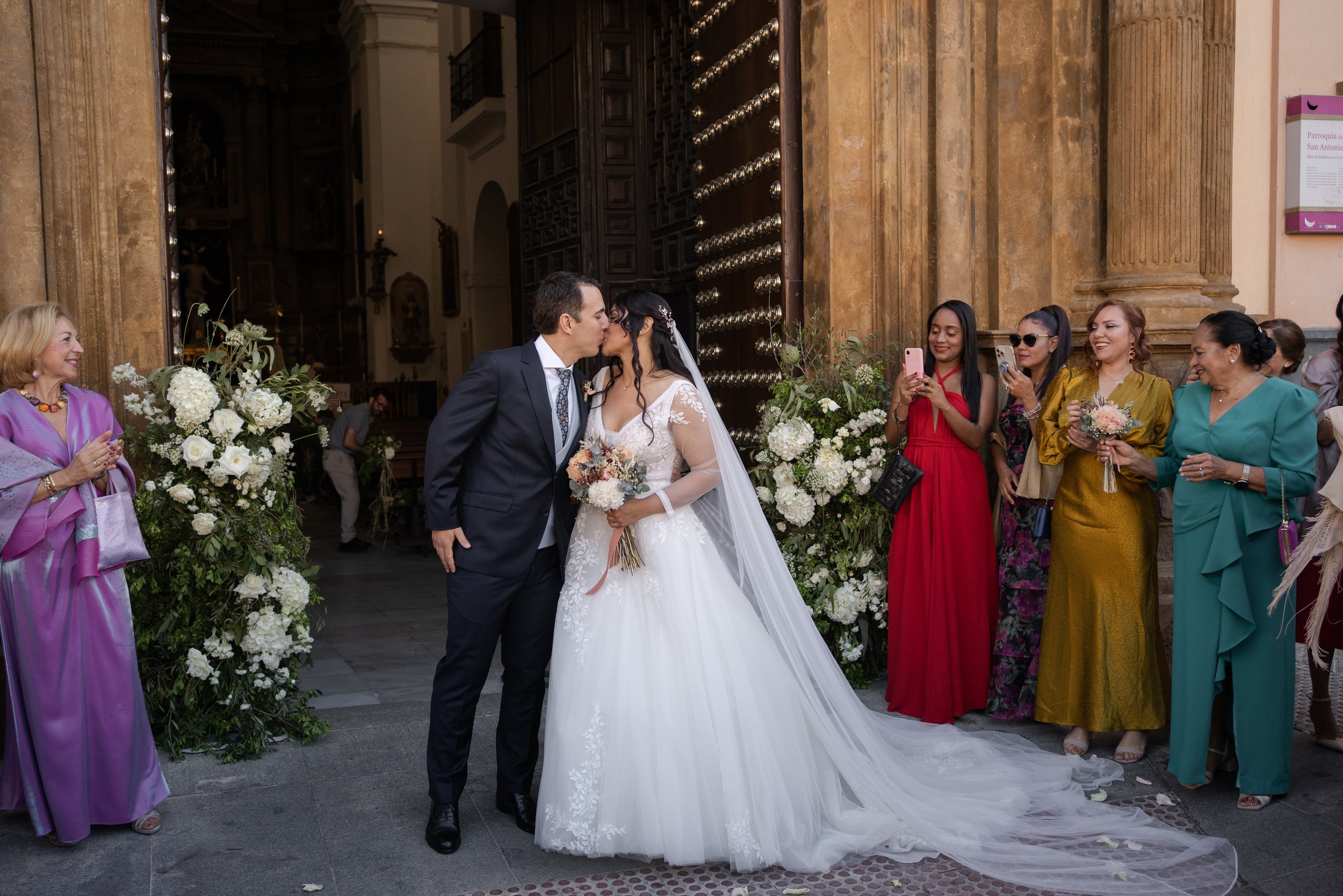 Pareja de recién casados besándose en la entrada de una iglesia en el casco historico de Cádiz, rodeados de invitados festivos y flores decorativas.