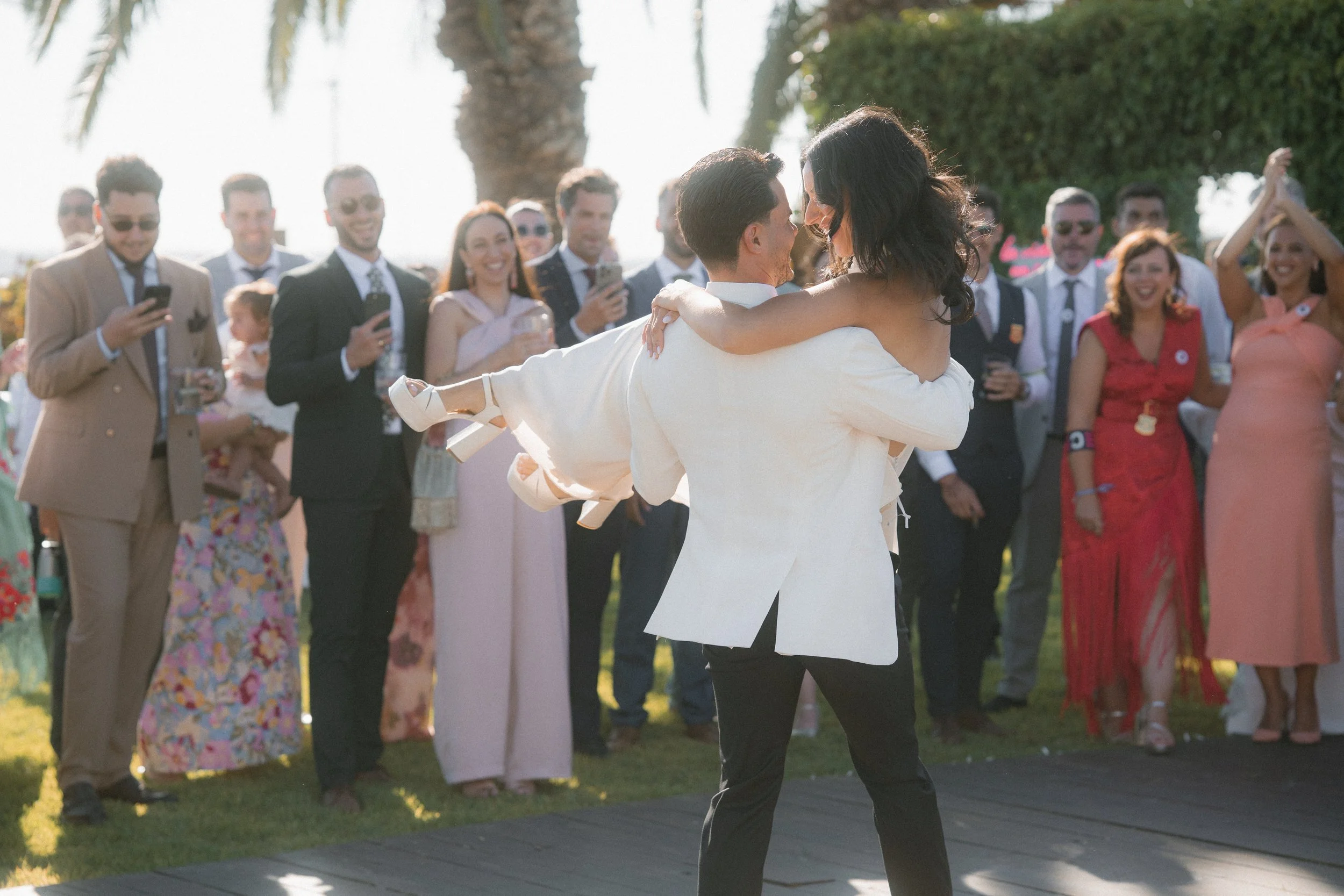 Una pareja bailando en una boda al aire libre, rodeada de invitados que toman fotografías y observan, en un ambiente luminoso y festivo con palmeras y árboles.
