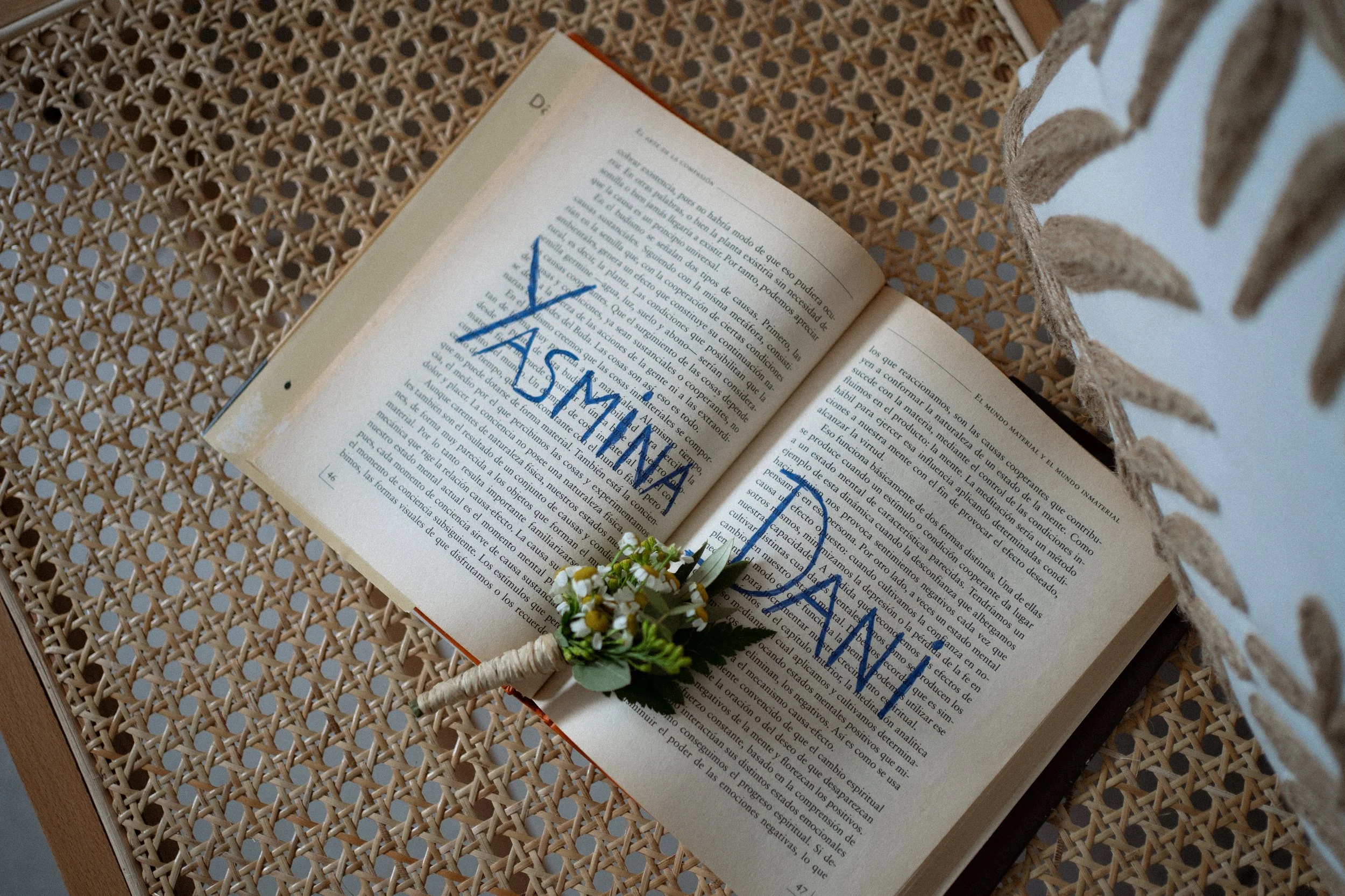 Libro abierto con tarjeta de felicitación sobre la mesa de mimbre, decorado con pequeña planta y “FELICIDADES” en letras azules en el libro.