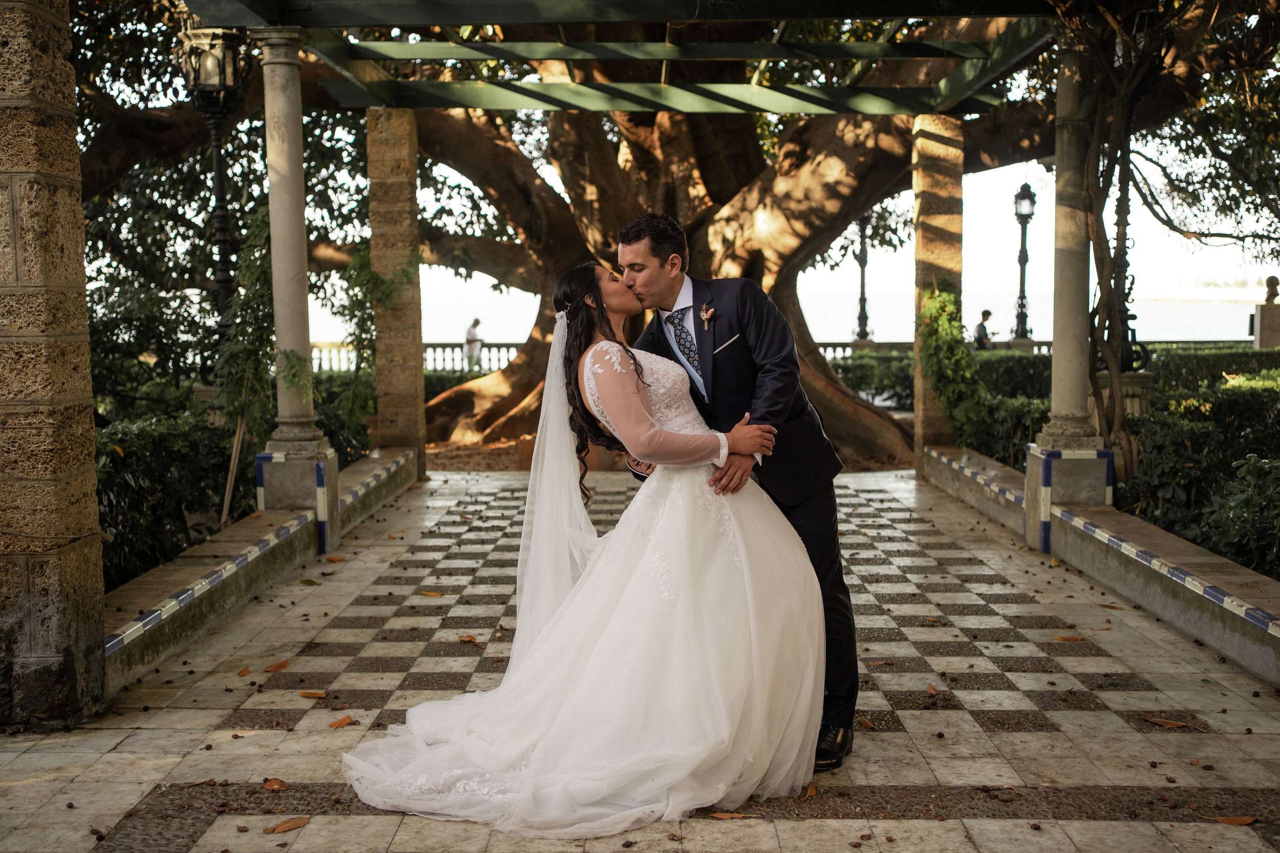Pareja de recién casados besándose en un entorno al aire libre con columnas de piedra y un gran árbol de fondo. Ella lleva un vestido de novia blanco y él un traje oscuro.