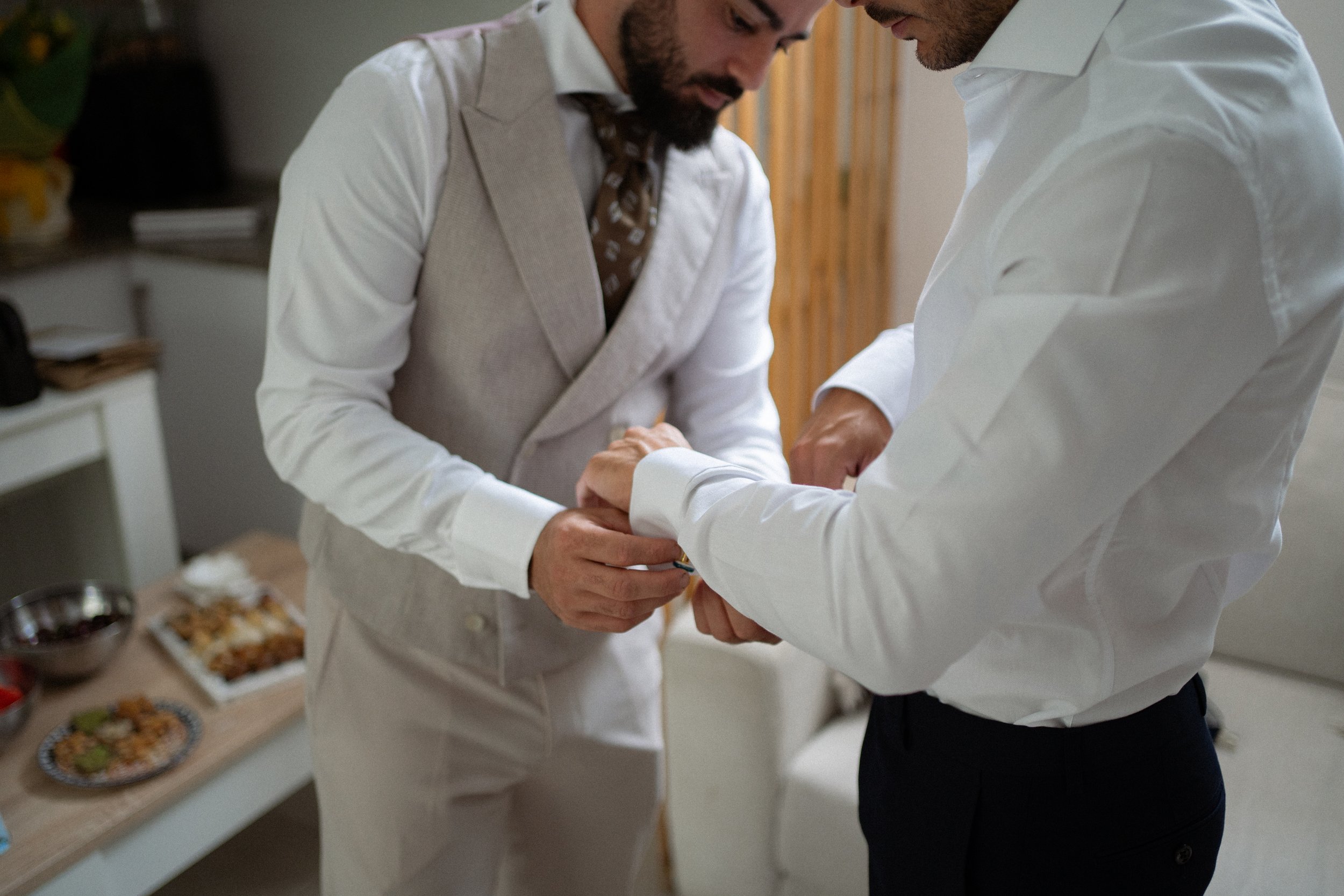 Novio y su hermano en ropa formal ajustándose las camisas en una sala con una mesa con comida en el fondo.