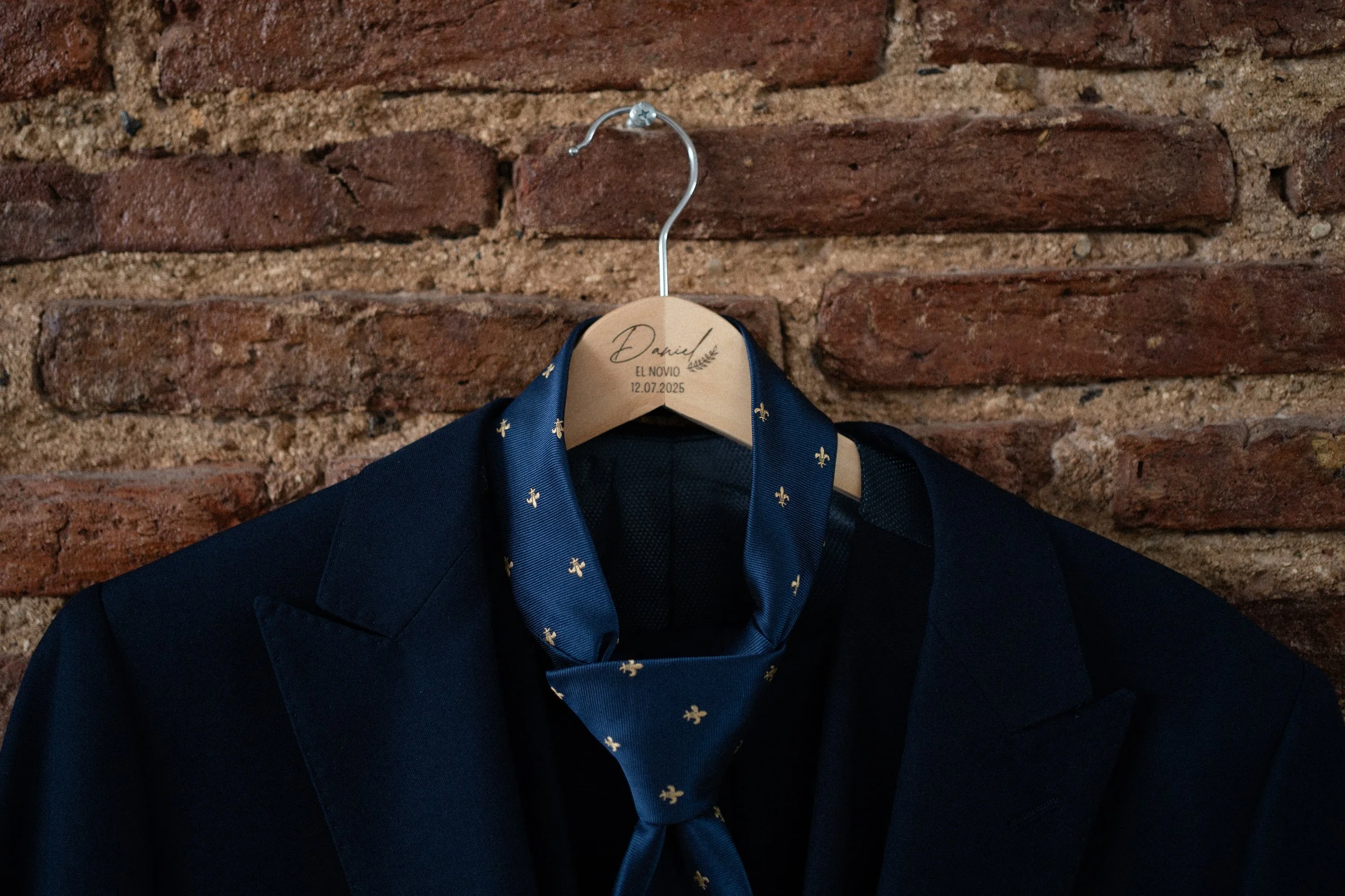 Un traje oscuro con corbata de seda azul con pequeños emblemas dorados, colgado en una percha de madera en una pared de ladrillos.