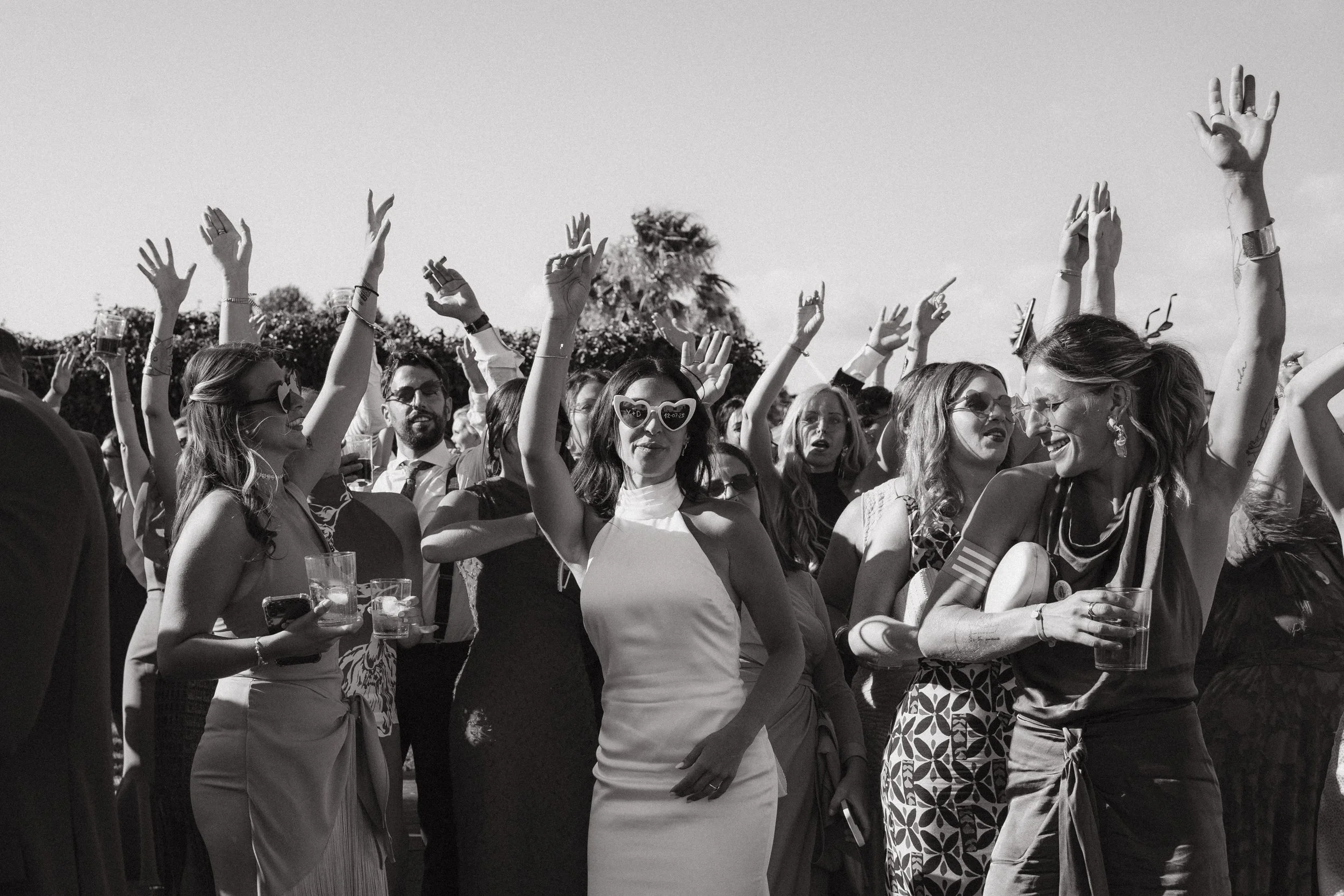 Personas en una fiesta, algunas con gafas de sol y vestidos elegantes, levantando las manos y sonriendo.