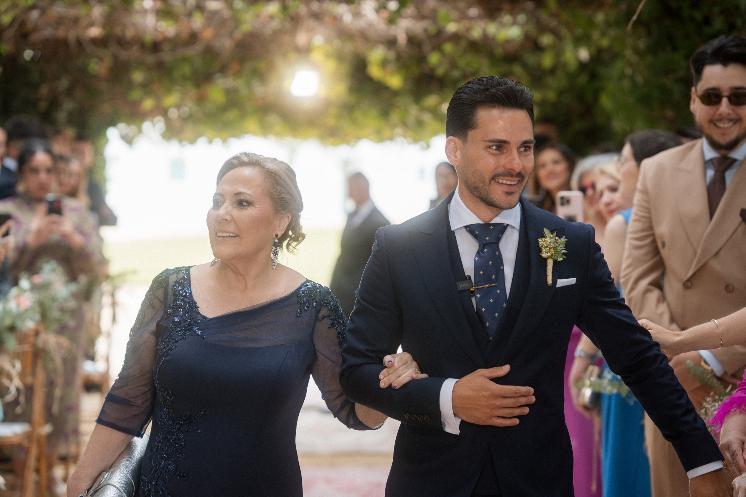 Hombre vestida con traje oscuro y mujer con vestido de noche en una ceremonia o boda al aire libre, rodeados de invitados.