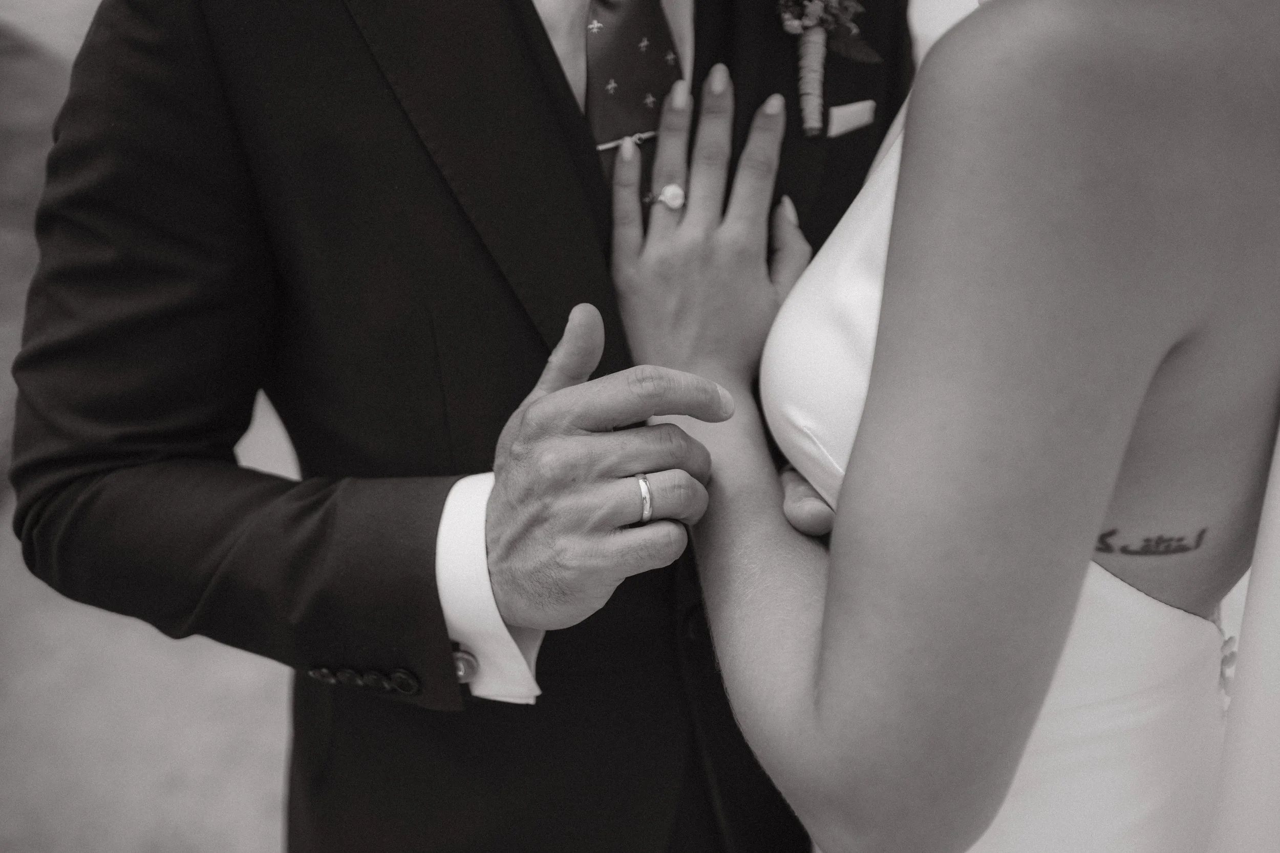 El novio con traje de etiqueta y una chica con vestido de novia sosteniéndose la mano izquierda con alianza en un momento de boda.