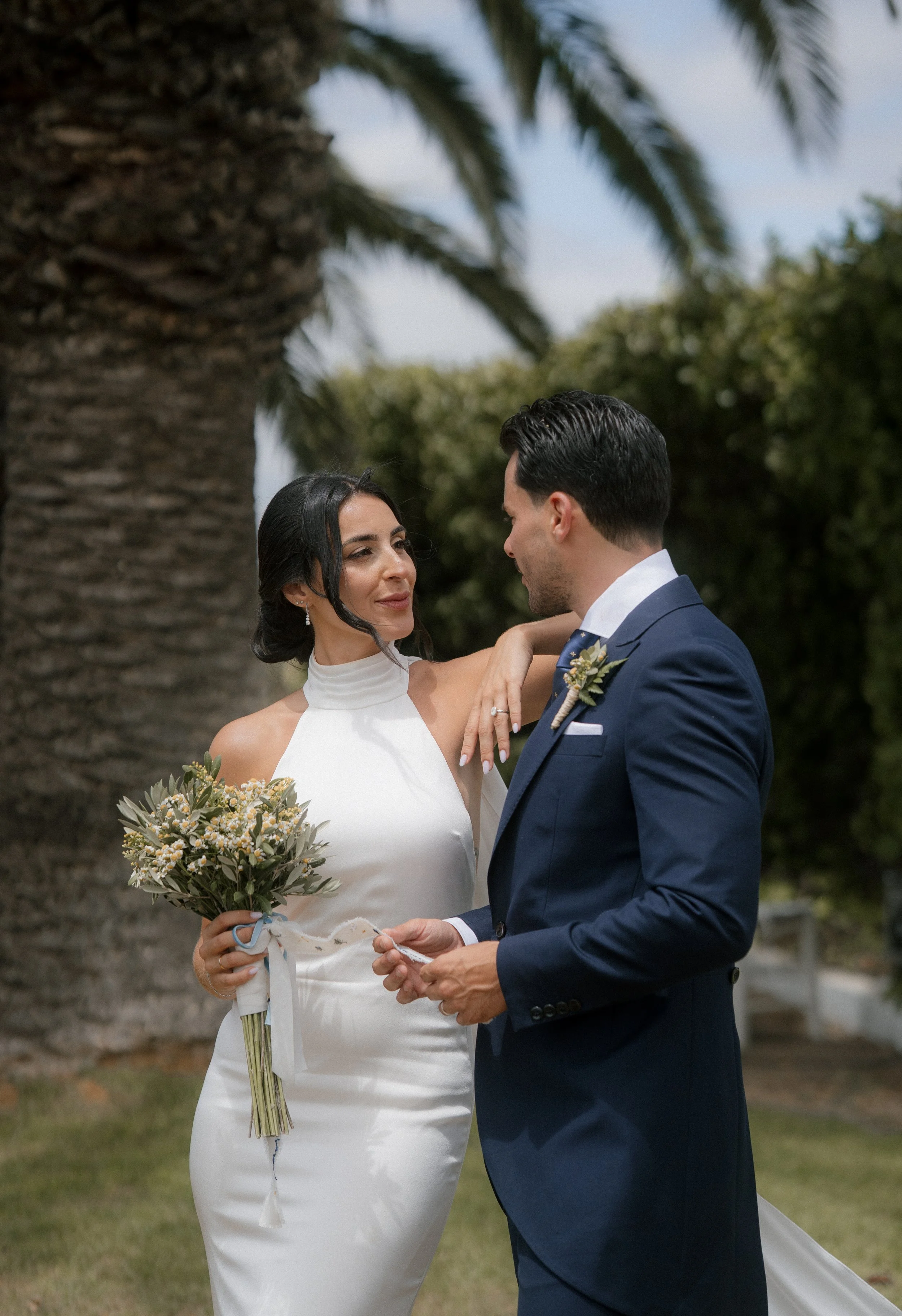 Una pareja de boda de pie frente a un árbol grande, la mujer con vestido blanco y el hombre con traje azul, mirándose amorosamente en un día soleado.