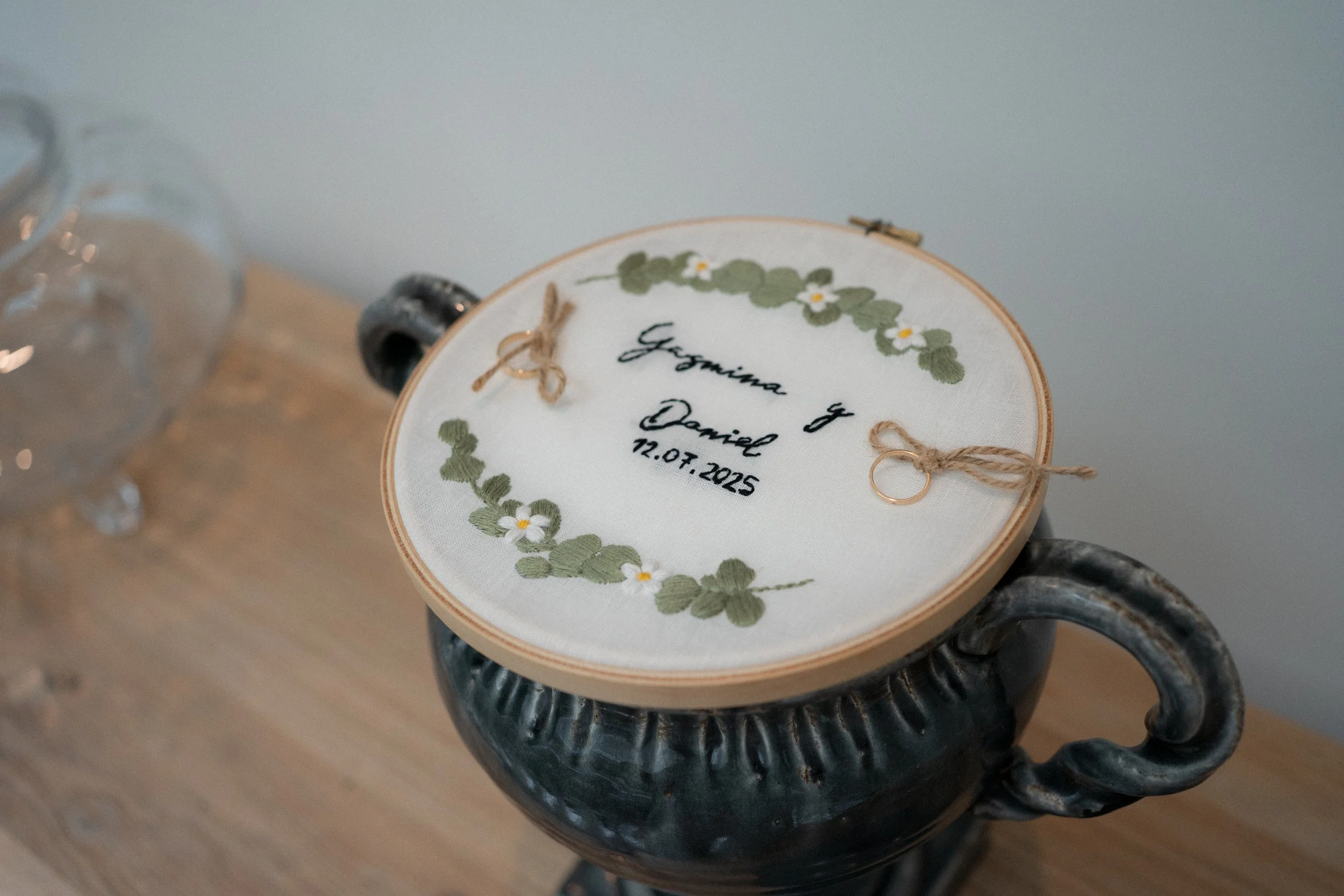 Una tela con bordado que tiene los nombres Yasmina y Daniel y la fecha 12.07.2025, decorada con flores y hojas en un círculo, colocada sobre una vasija de cerámica negra.