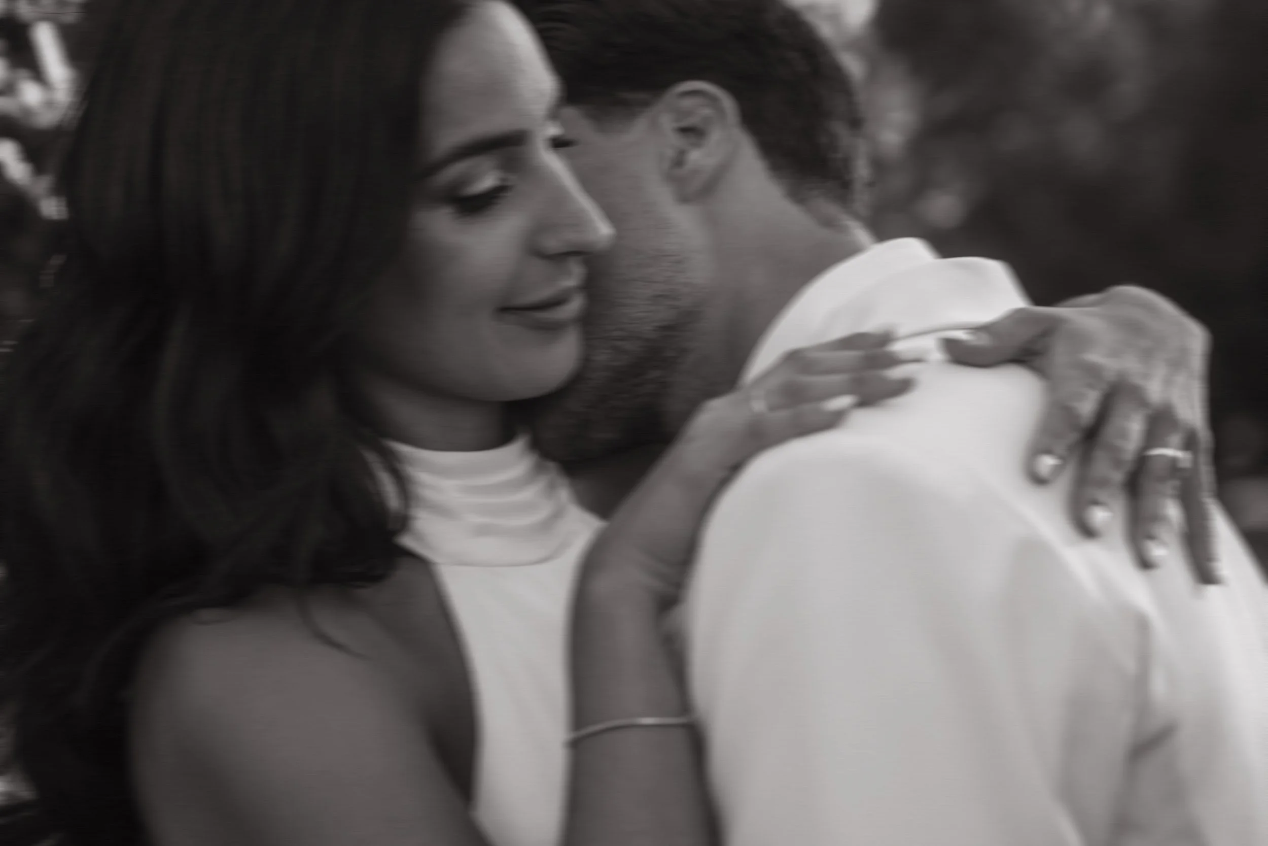 Pareja en un abrazo cercano, ella con ojos cerrados y sonriendo, él con la cabeza inclinada hacia ella, en un fondo difuso. Imagen en blanco y negro.