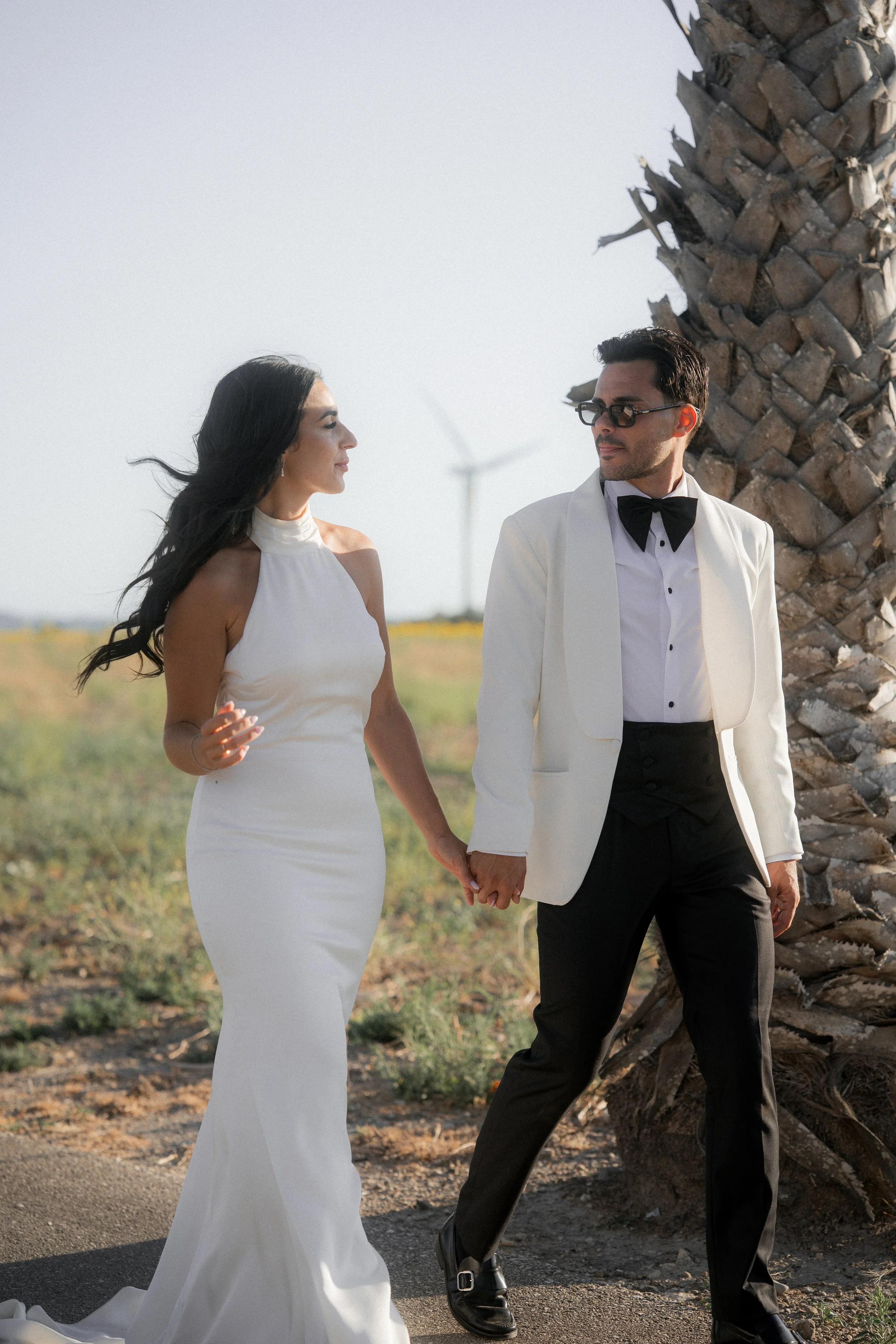 Pareja vestida formalmente, la mujer en vestido blanco y el hombre con saco y pajarita, caminando juntos cerca de una palmera en un campo con un aerogenerador de fondo.