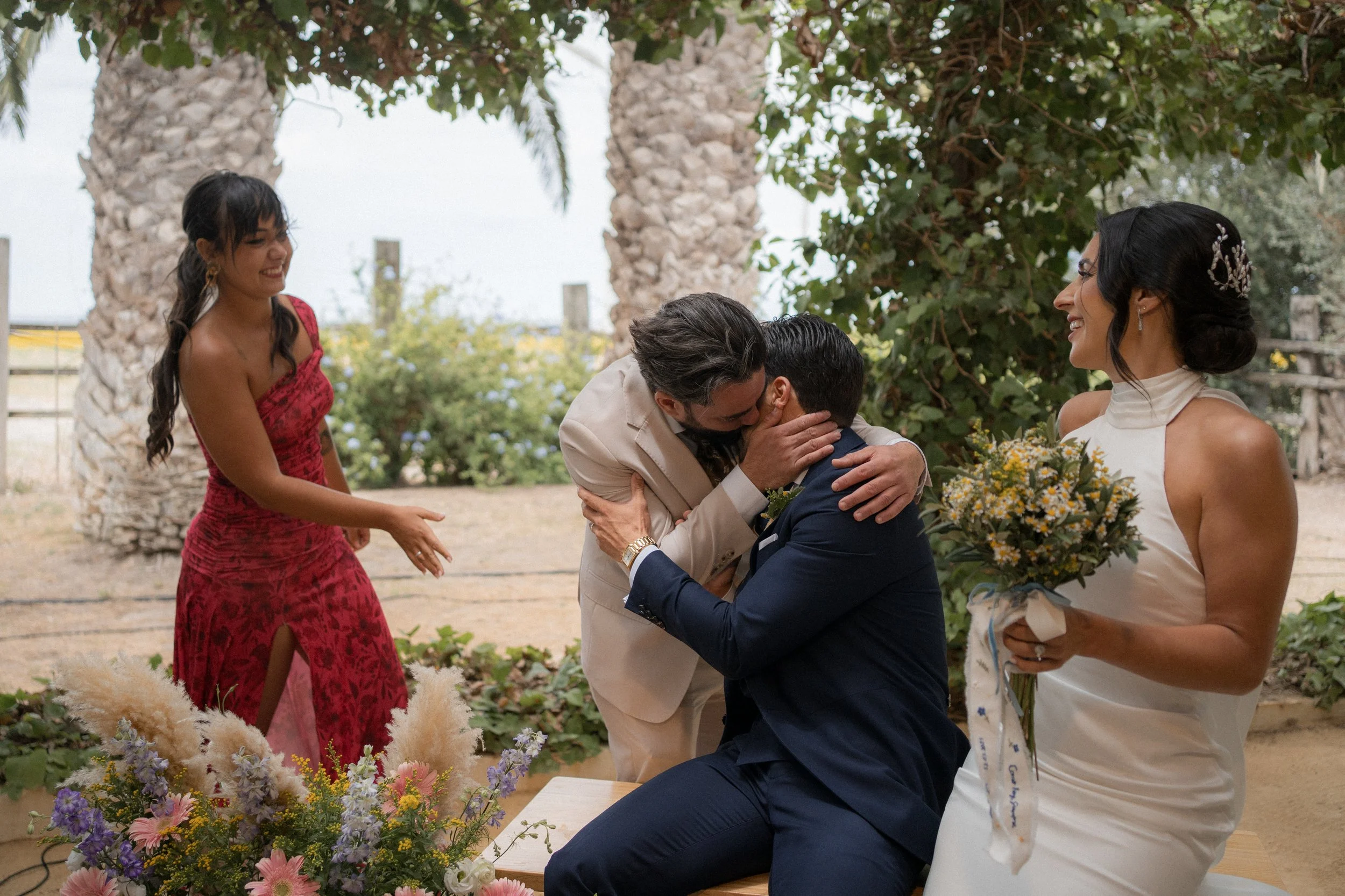 Pareja de hombres en el momento de un beso en una boda, rodeados de tres mujeres, una de ellas con un ramo de flores, en un lugar al aire libre con árboles y plantas.