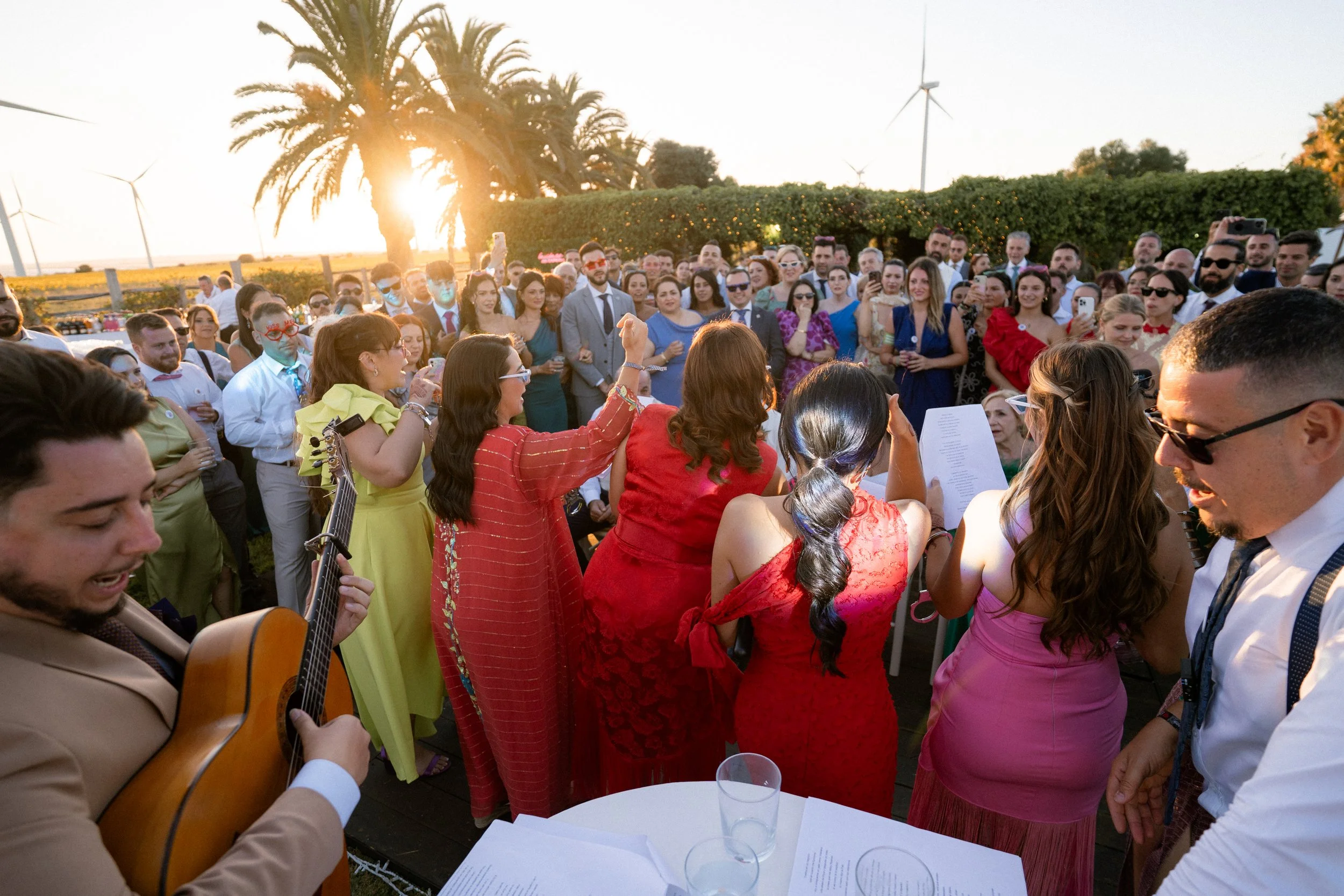 Celebración al aire libre con grupo de personas cantando cuplés, algunos con gafas de sol, en un evento cerca del atardecer, con palmeras y molinos de viento al fondo, en un ambiente festivo y social.