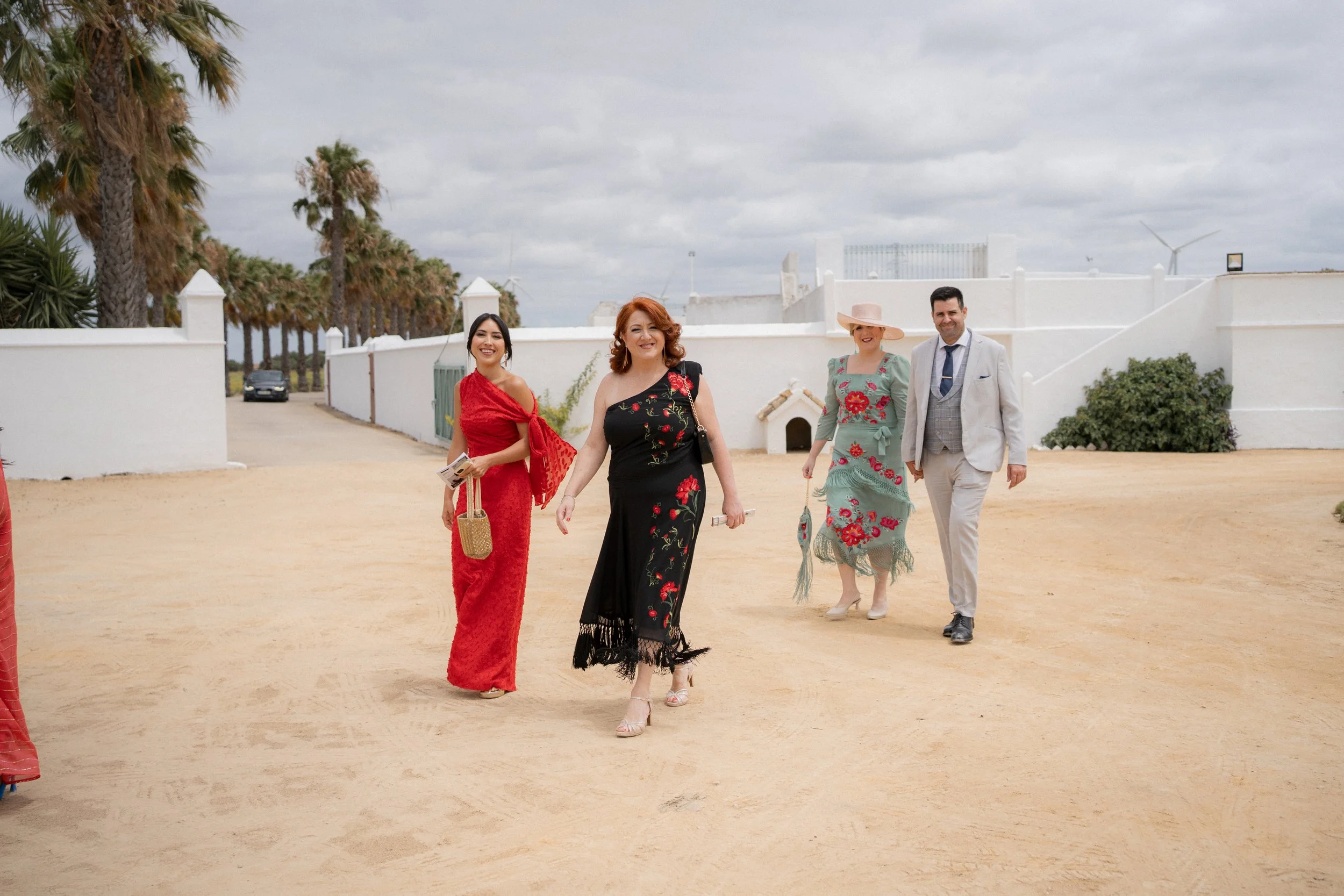 Cuatro personas caminando en un camino de tierra con fondo de palmeras, paredes blancas y cielo nublado.